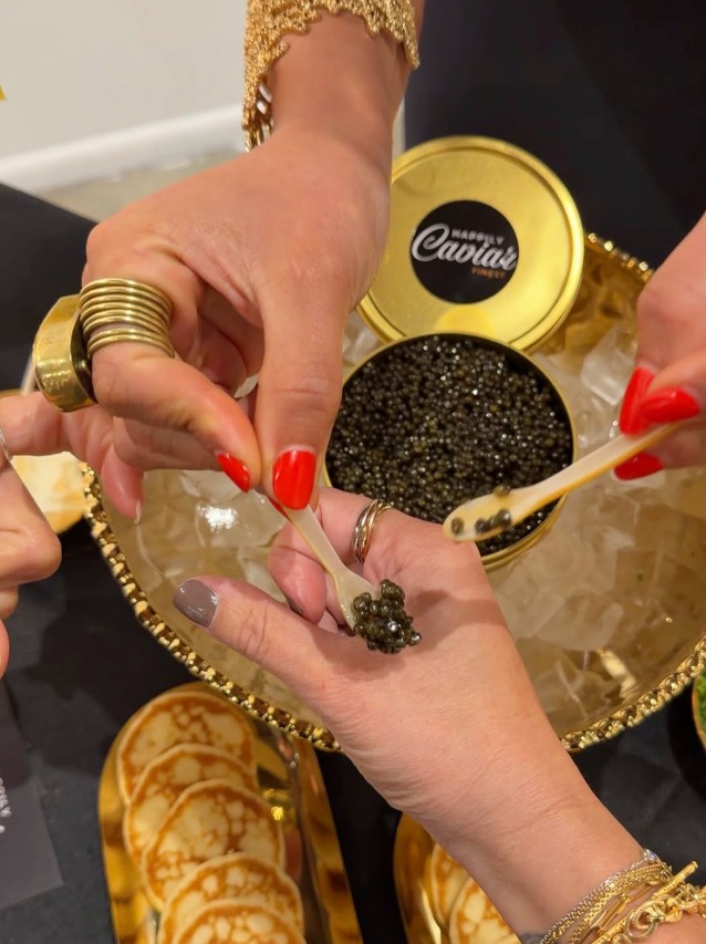 Two hands exchanging black caviar with a small spoon, gold jewelry, and a gold container in the background.