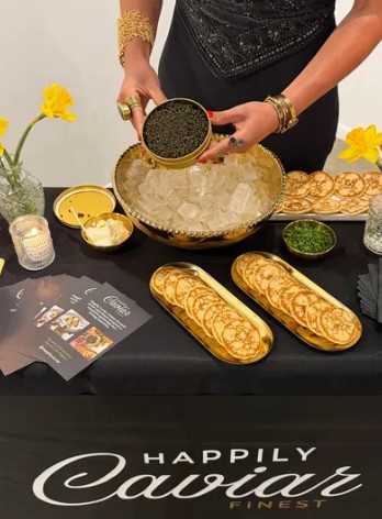 Person in black dress holding a round container over a table with gold jewelry, yellow flowers, and decorative items.