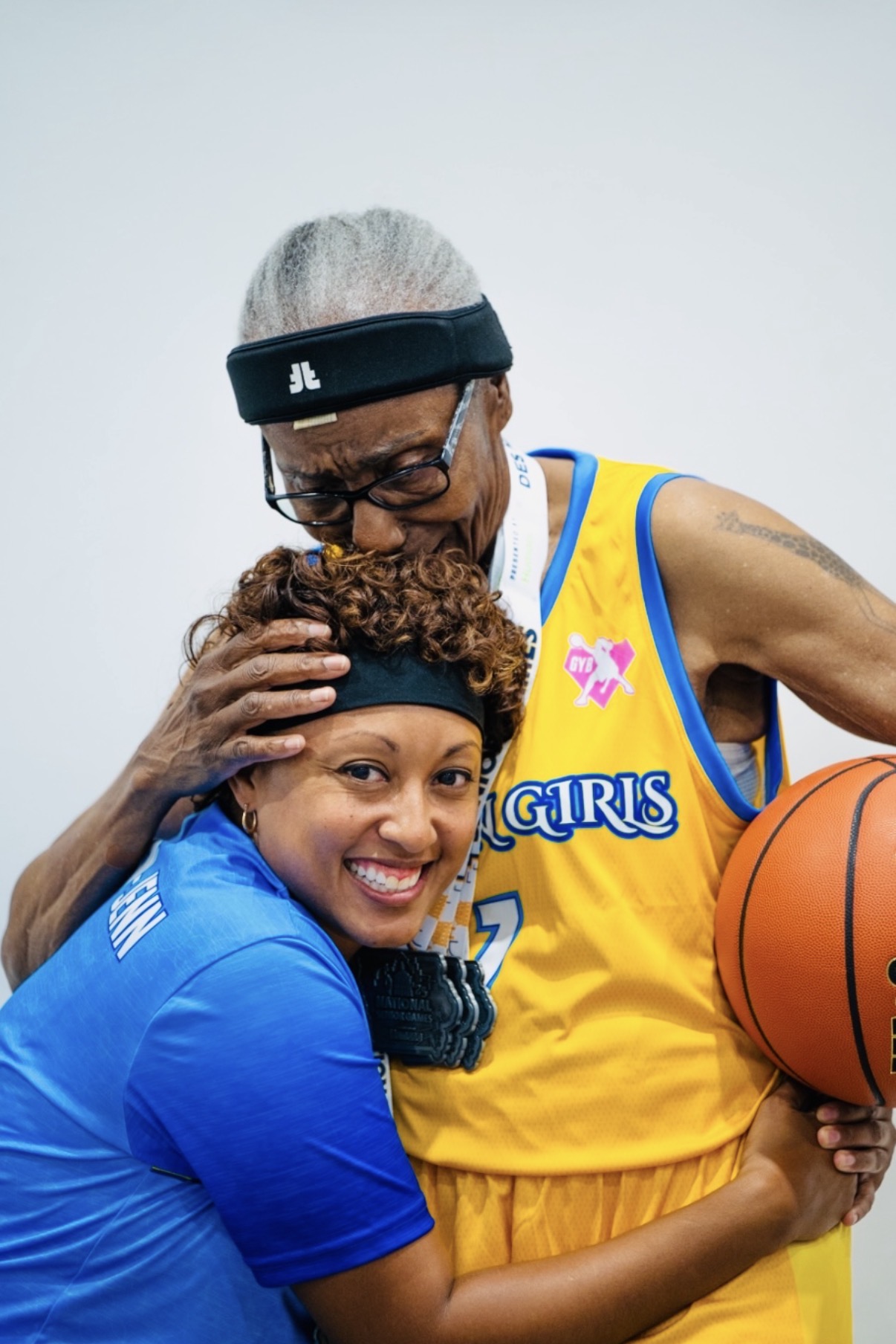 Two women, one in a blue shirt and the other in a yellow basketball uniform, smiling and holding a basketball.