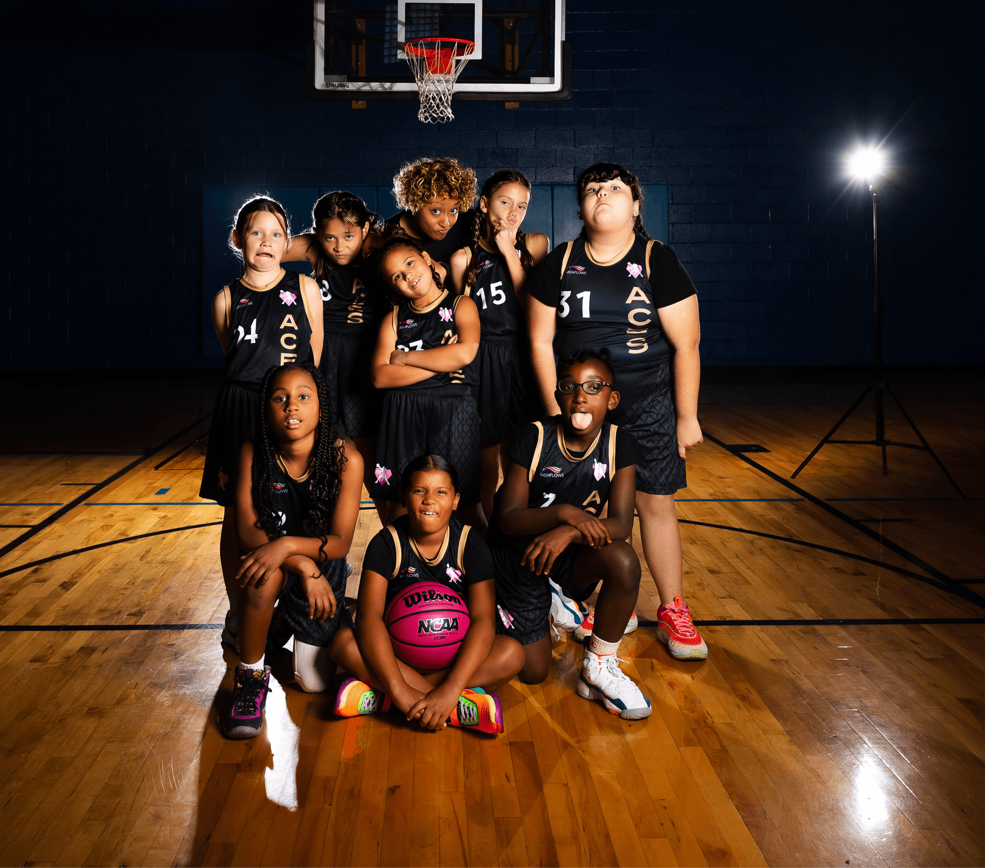 Group of young female basketball players in black uniforms posing on a court with a basketball hoop behind them.