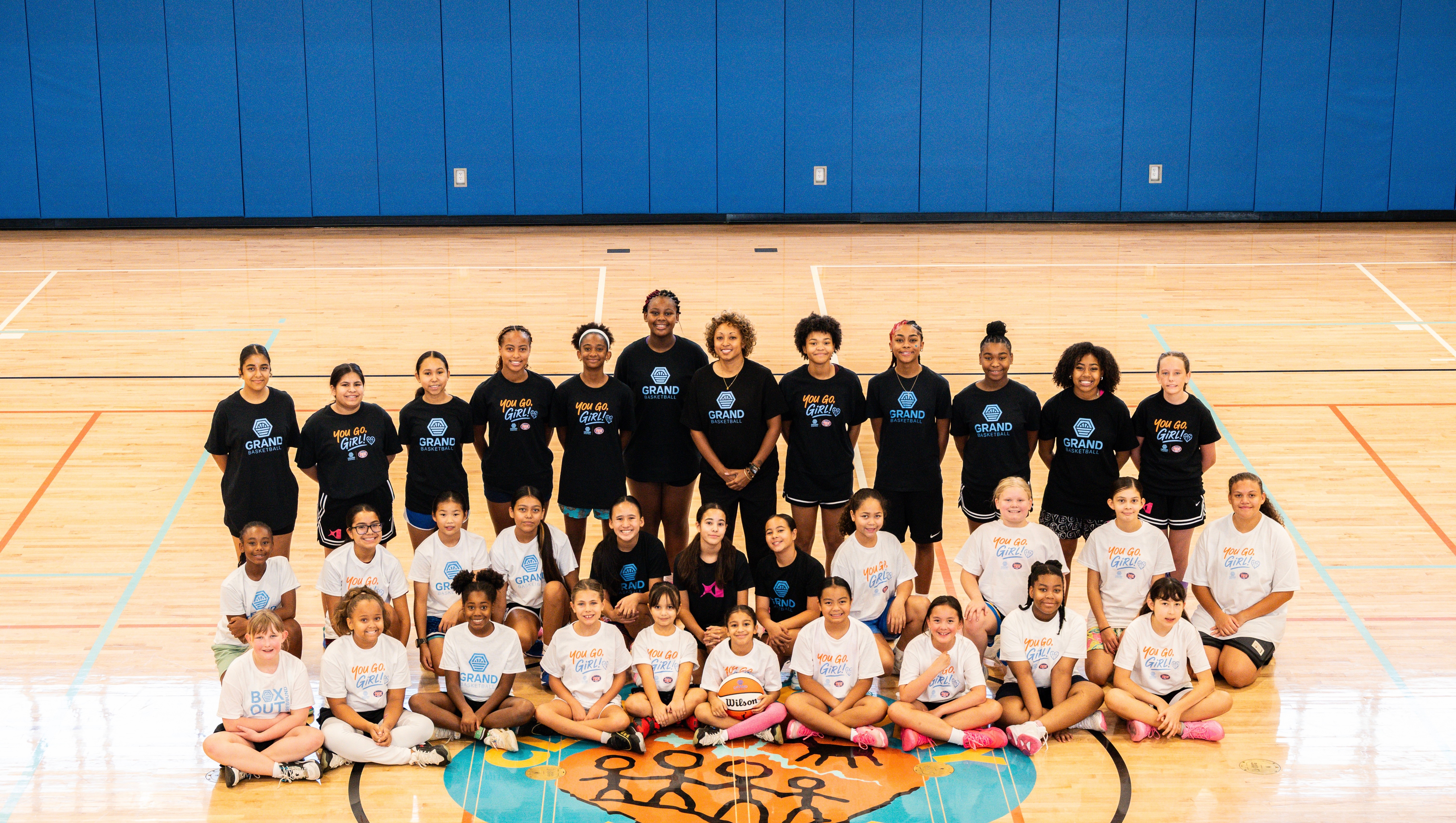 Group of children and adults in a gymnasium, posing for a team photo on a basketball court, with a blue wall in the background.