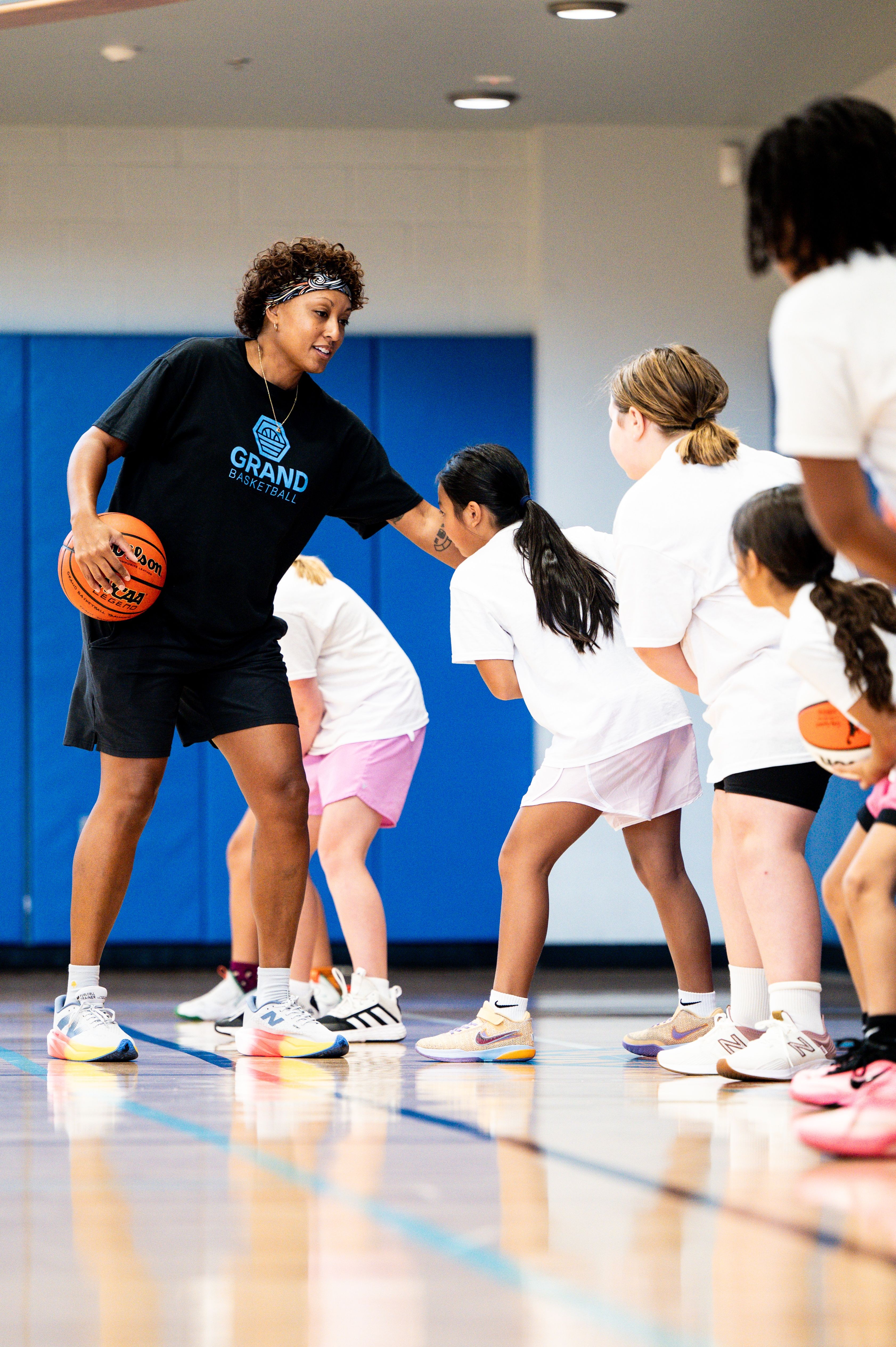 Coach with basketball instructs young girls in a gym, all wearing white shirts and shorts, some holding basketballs.