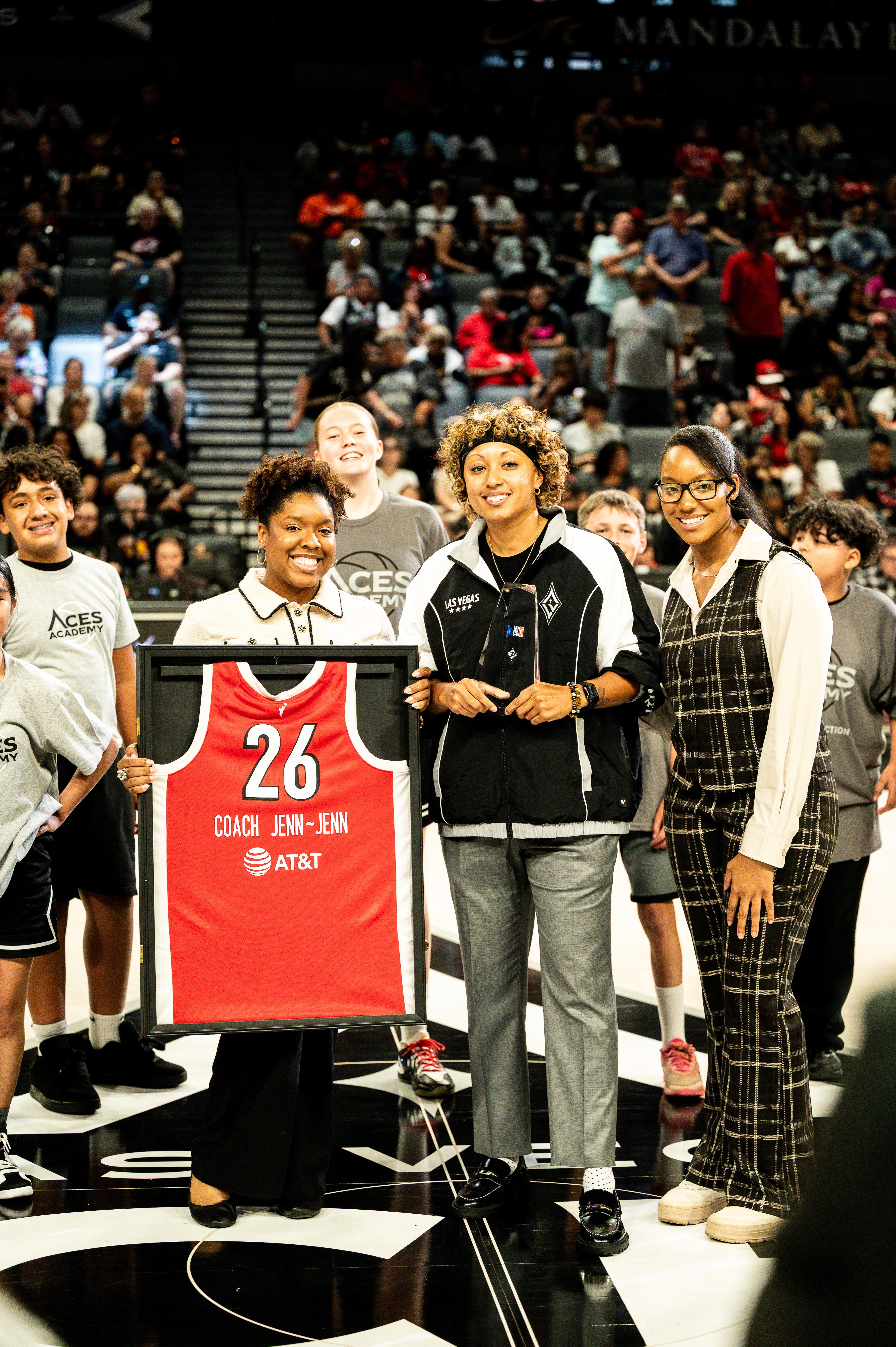 Group of people standing on a basketball court, holding a red basketball jersey, with spectators in the background.
