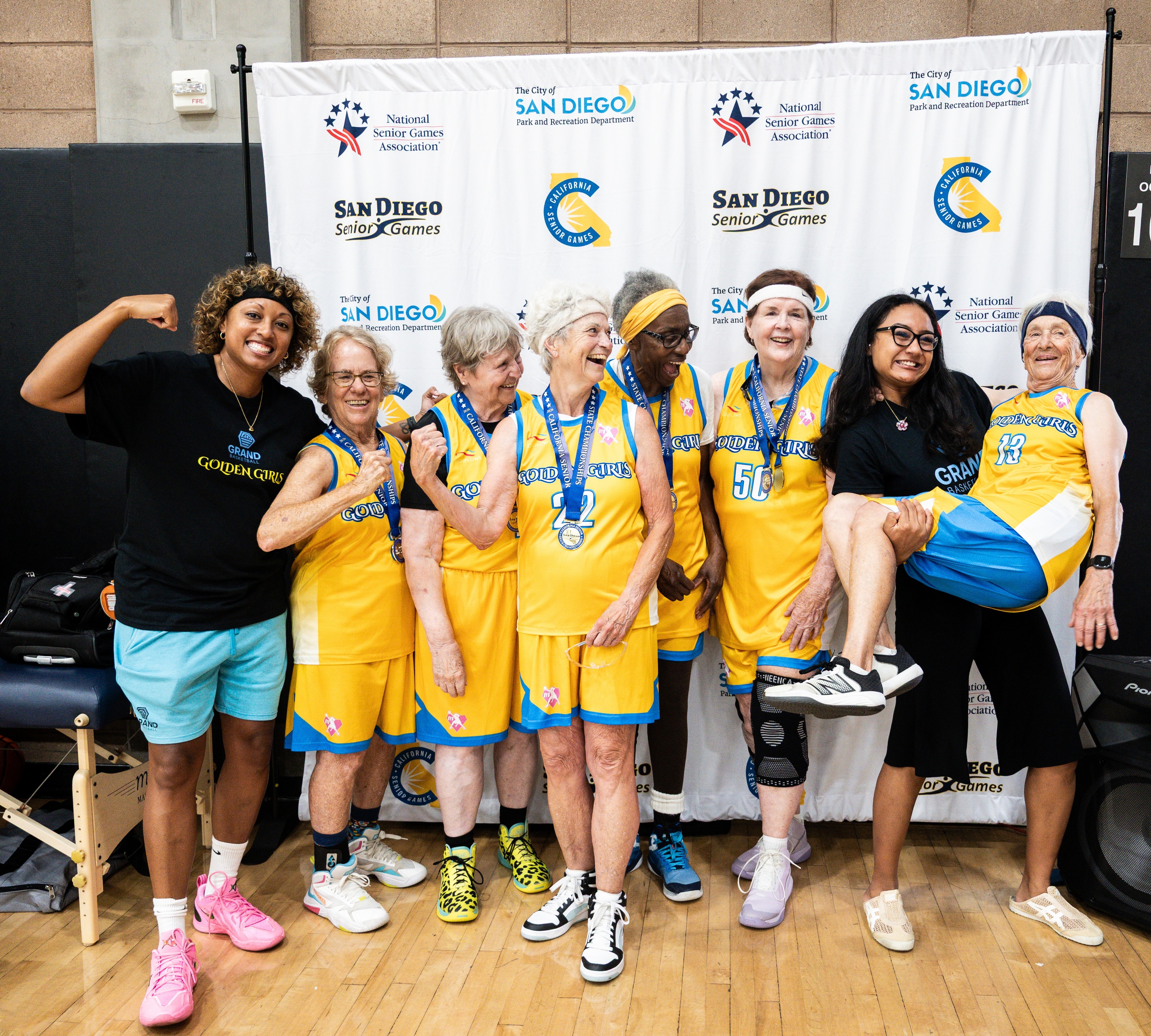 Group of women and a girl in yellow and blue sports uniforms, some wearing medals, posing indoors in front of a backdrop.