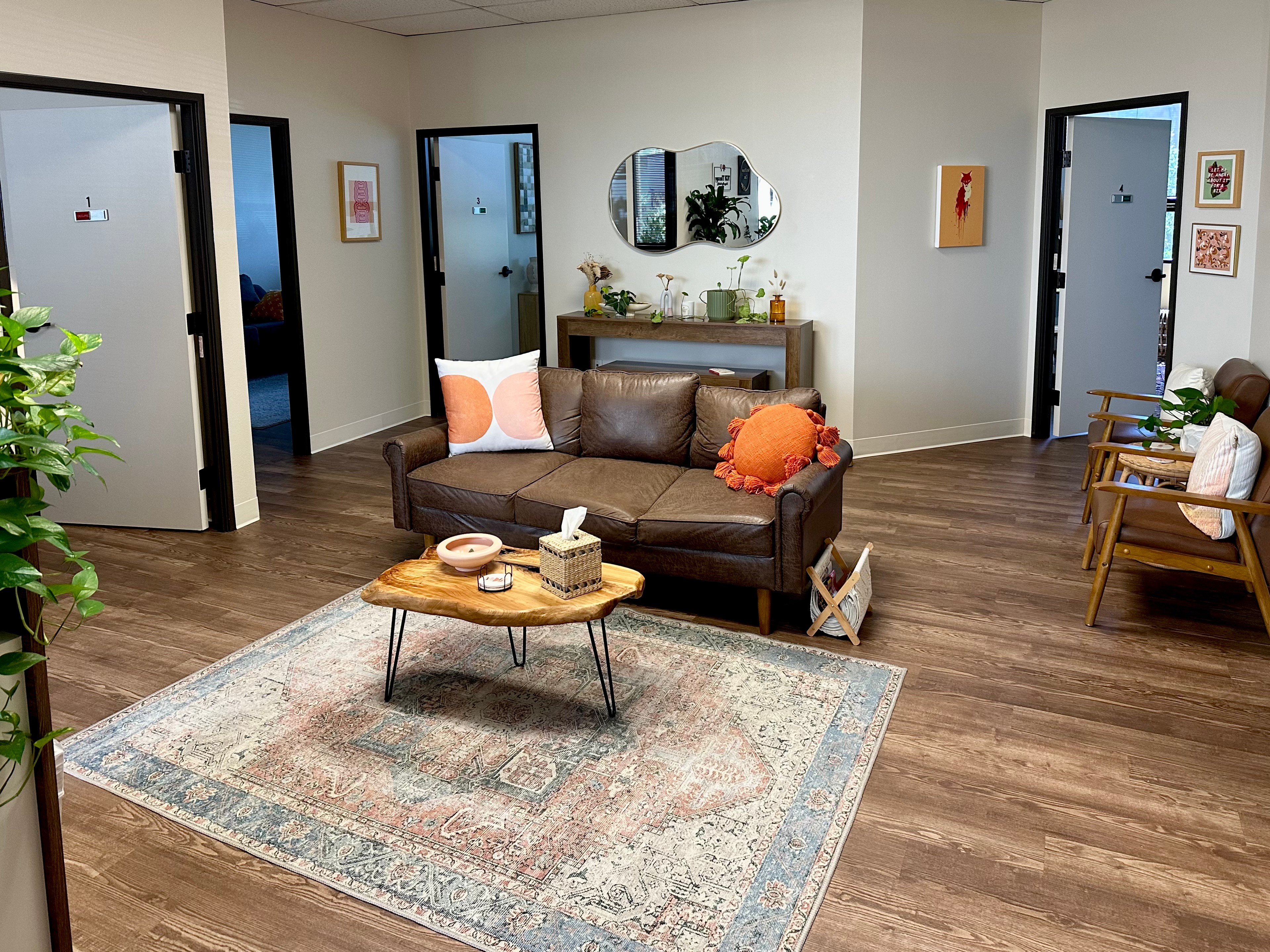 Living room with a brown sofa, wooden coffee table, and a patterned rug. Walls have framed art and mirrors, with doors leading to other rooms.