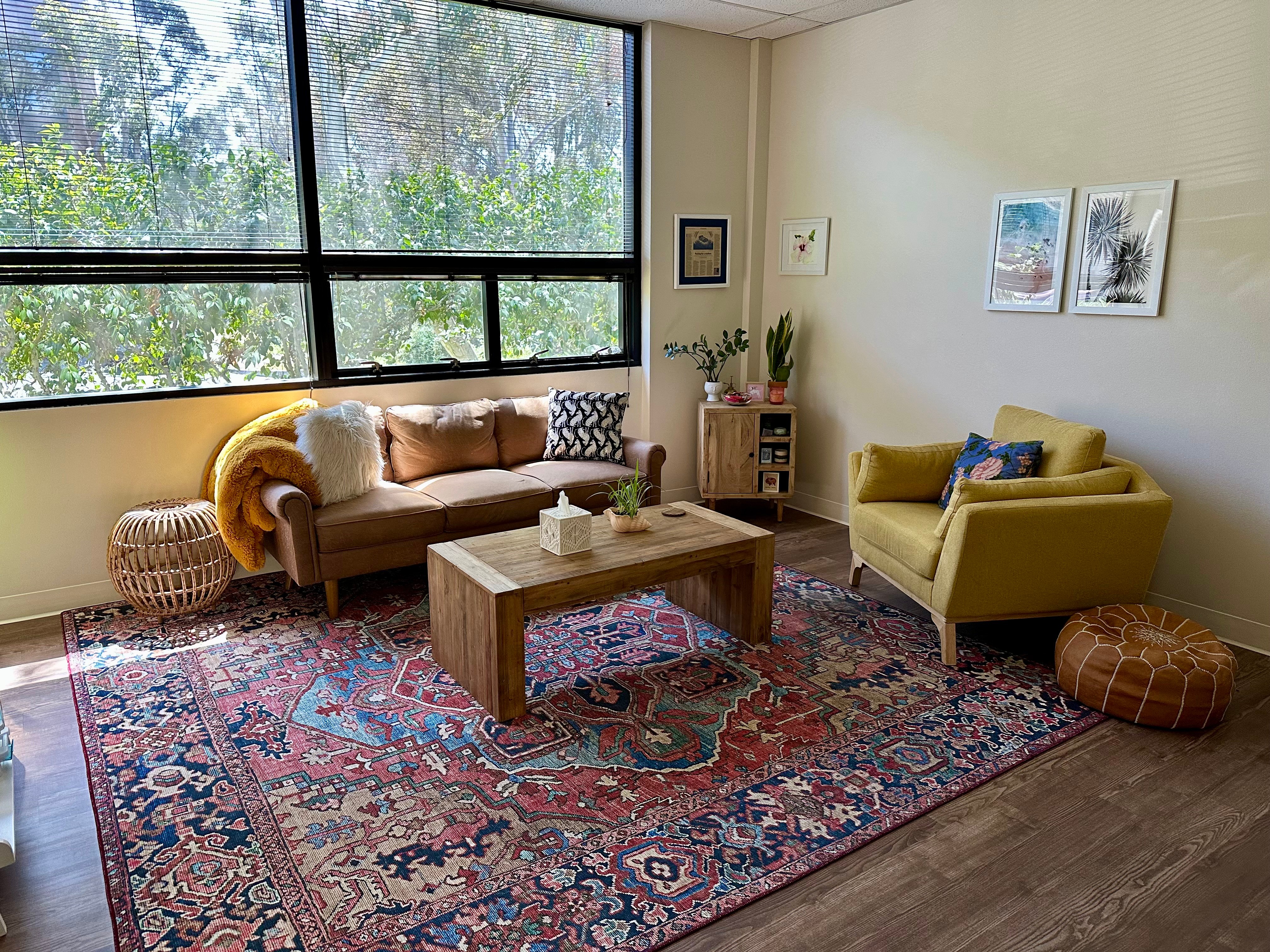 Living room with a large window, sofa, armchair, wooden coffee table, and patterned rug, decorated with framed pictures and plants.