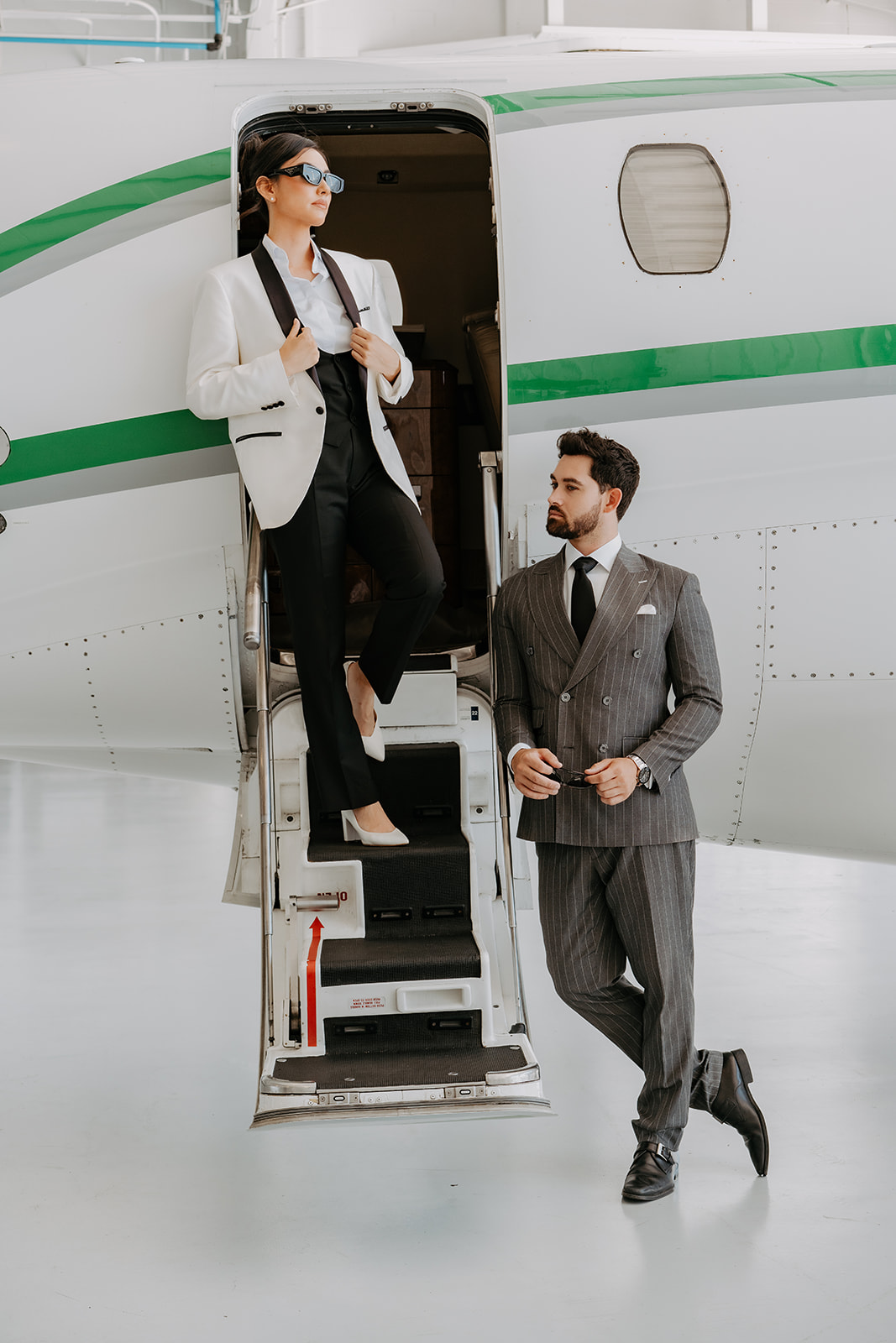 A woman in a white blazer and black pants stands on airplane stairs, talking to a man in a gray suit outside.