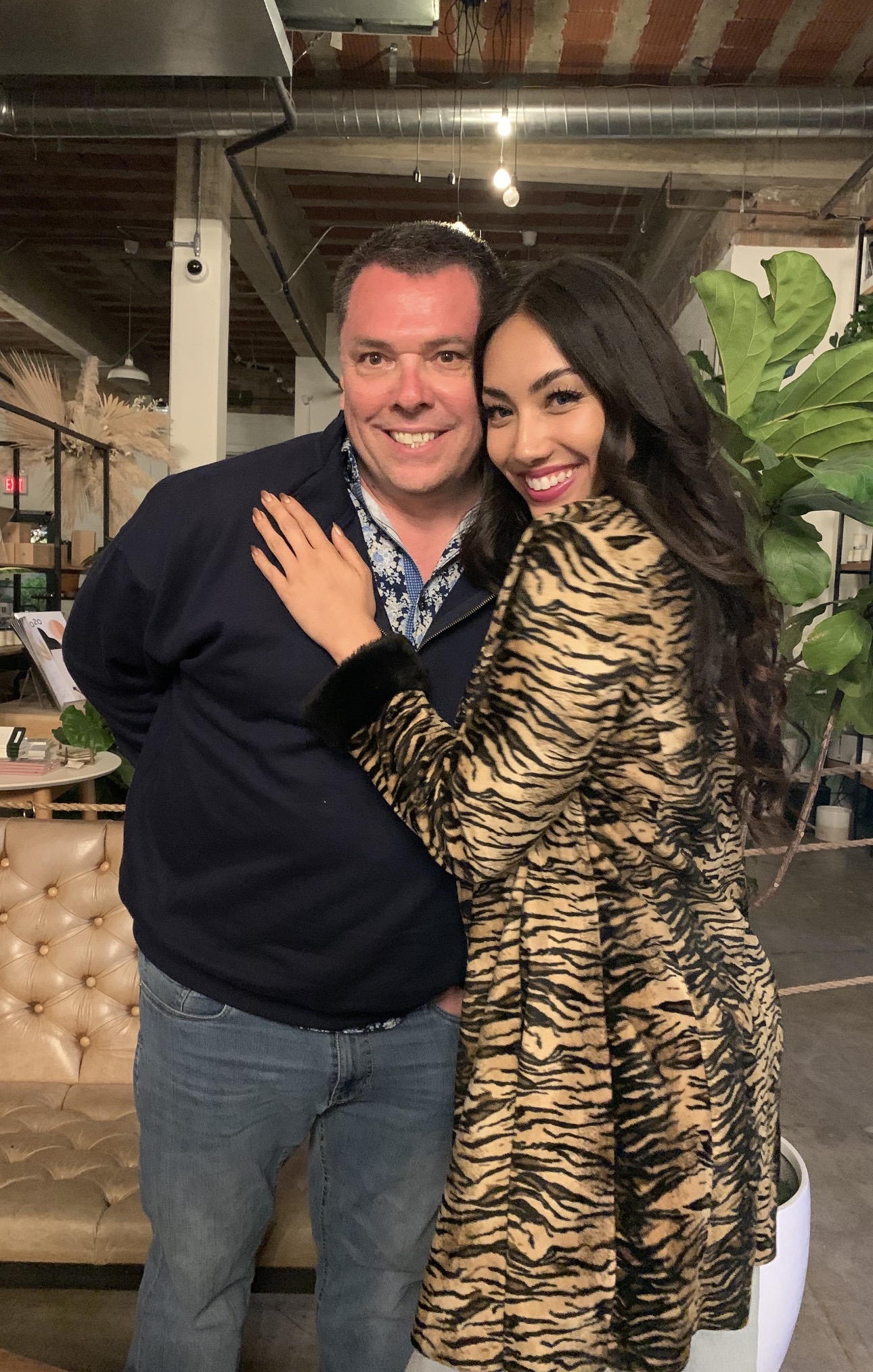 Two people smiling and hugging in an indoor setting with plants and furniture visible in the background.