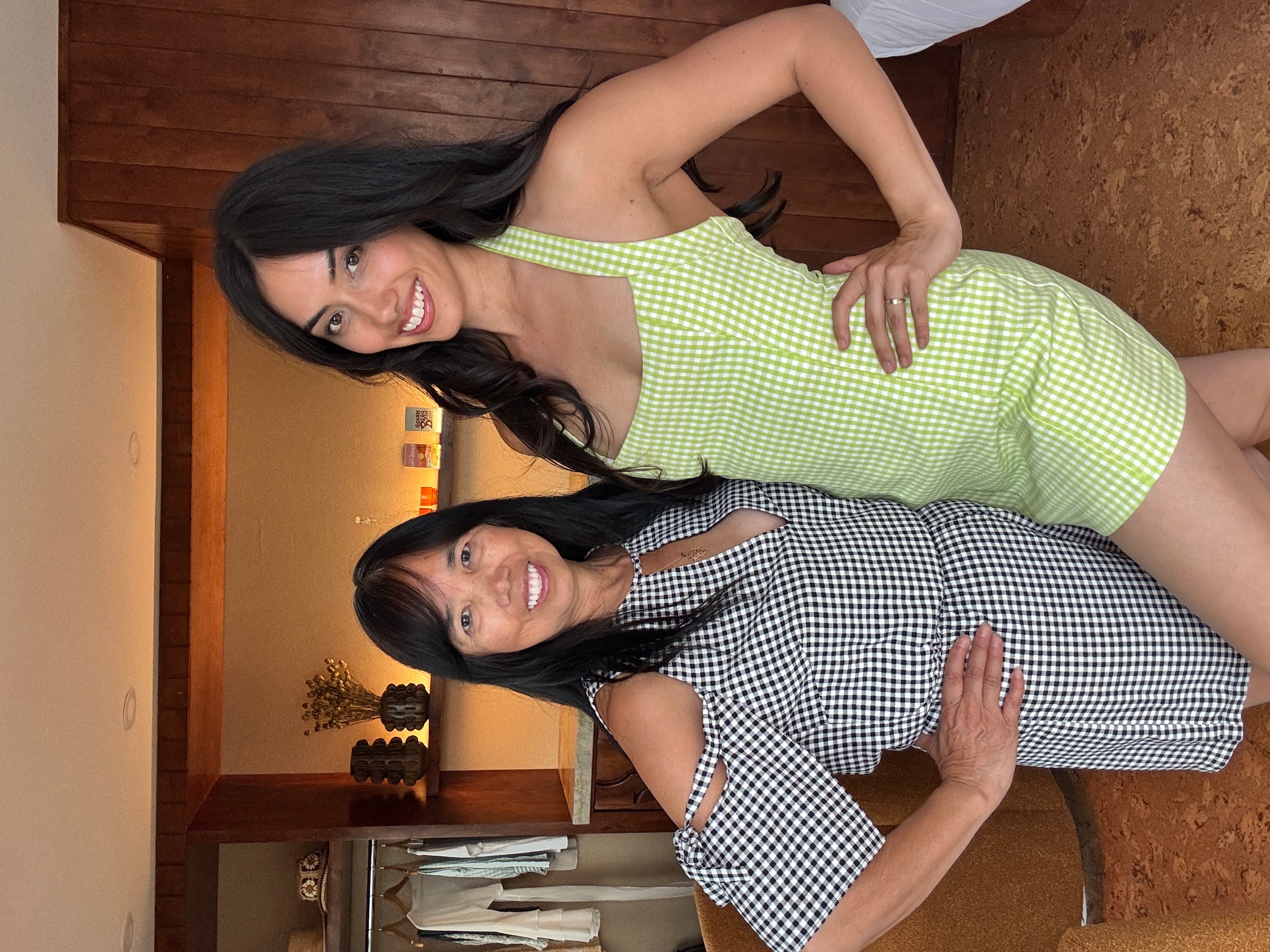 Two women standing indoors, one in a green checkered dress and the other in a black and white checkered dress, smiling.