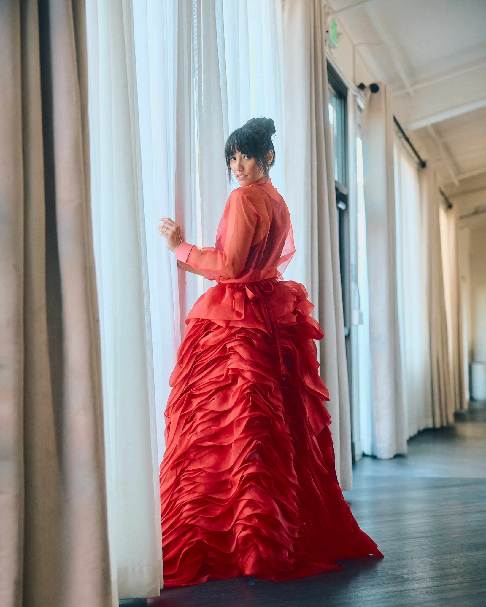 Woman in a red, ruffled gown standing by a window with white curtains, looking over her shoulder inside a bright room.