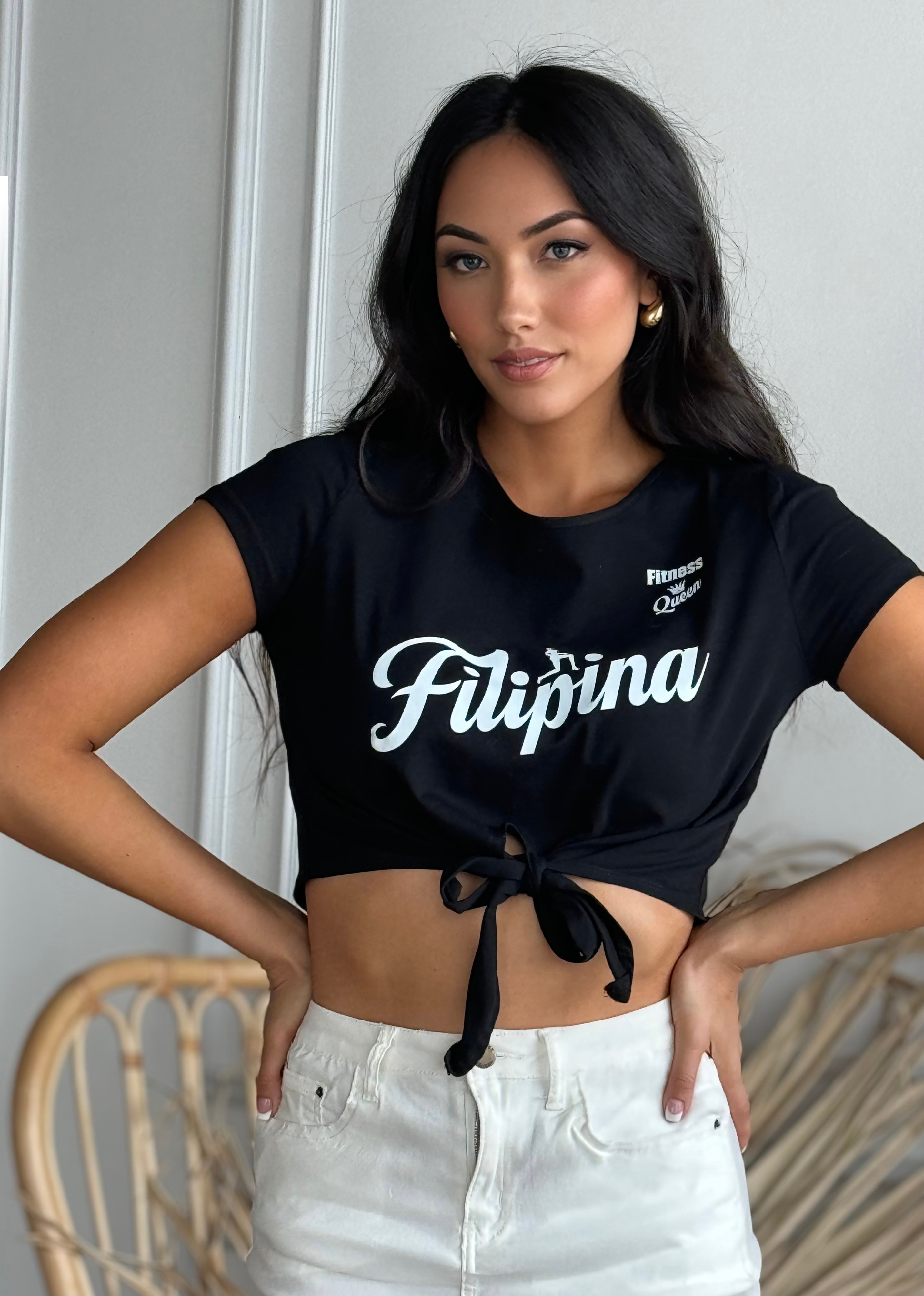 Young woman with long dark hair wearing a black crop top tied at the waist and white pants, standing indoors.