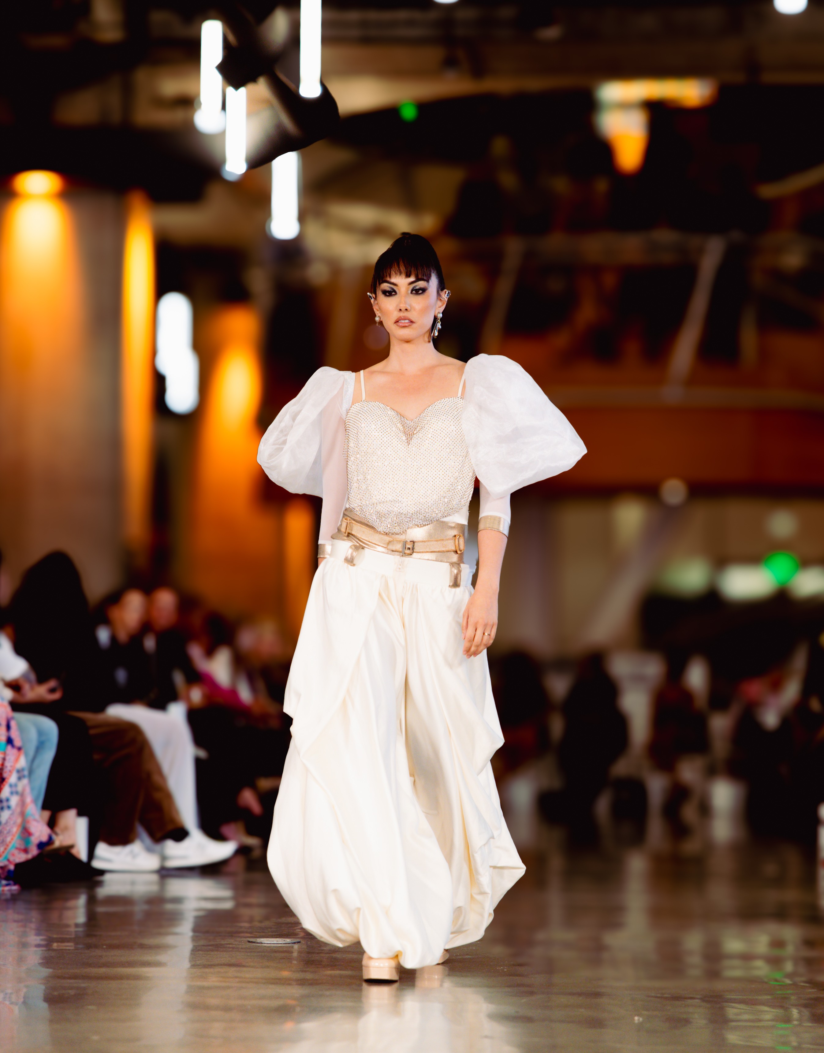 Woman walking on runway in white dress with puffed sleeves, in a large indoor event space with audience.