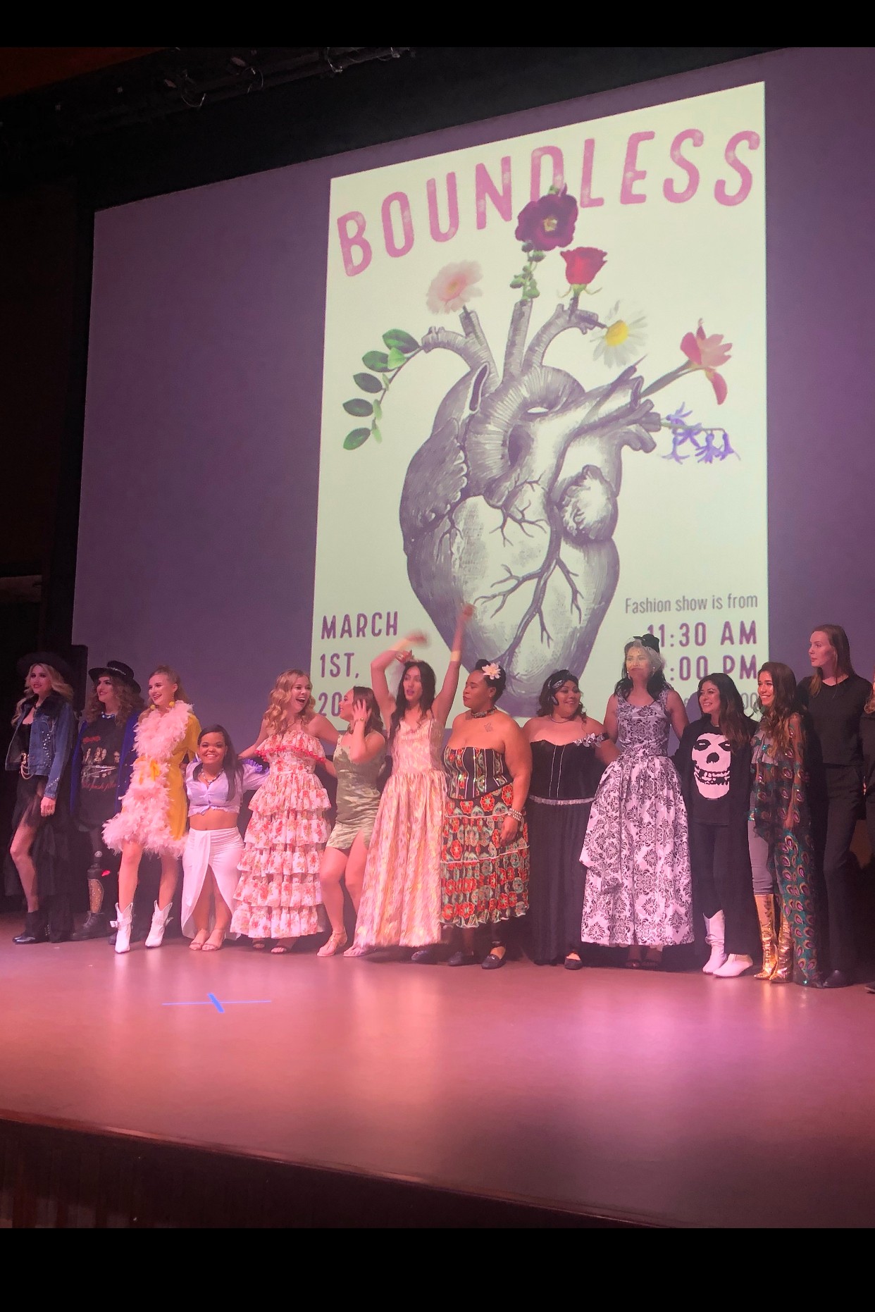 Group of people in colorful costumes on stage with a large poster of a heart with flowers and the word 'BOUNDLESS' behind them.