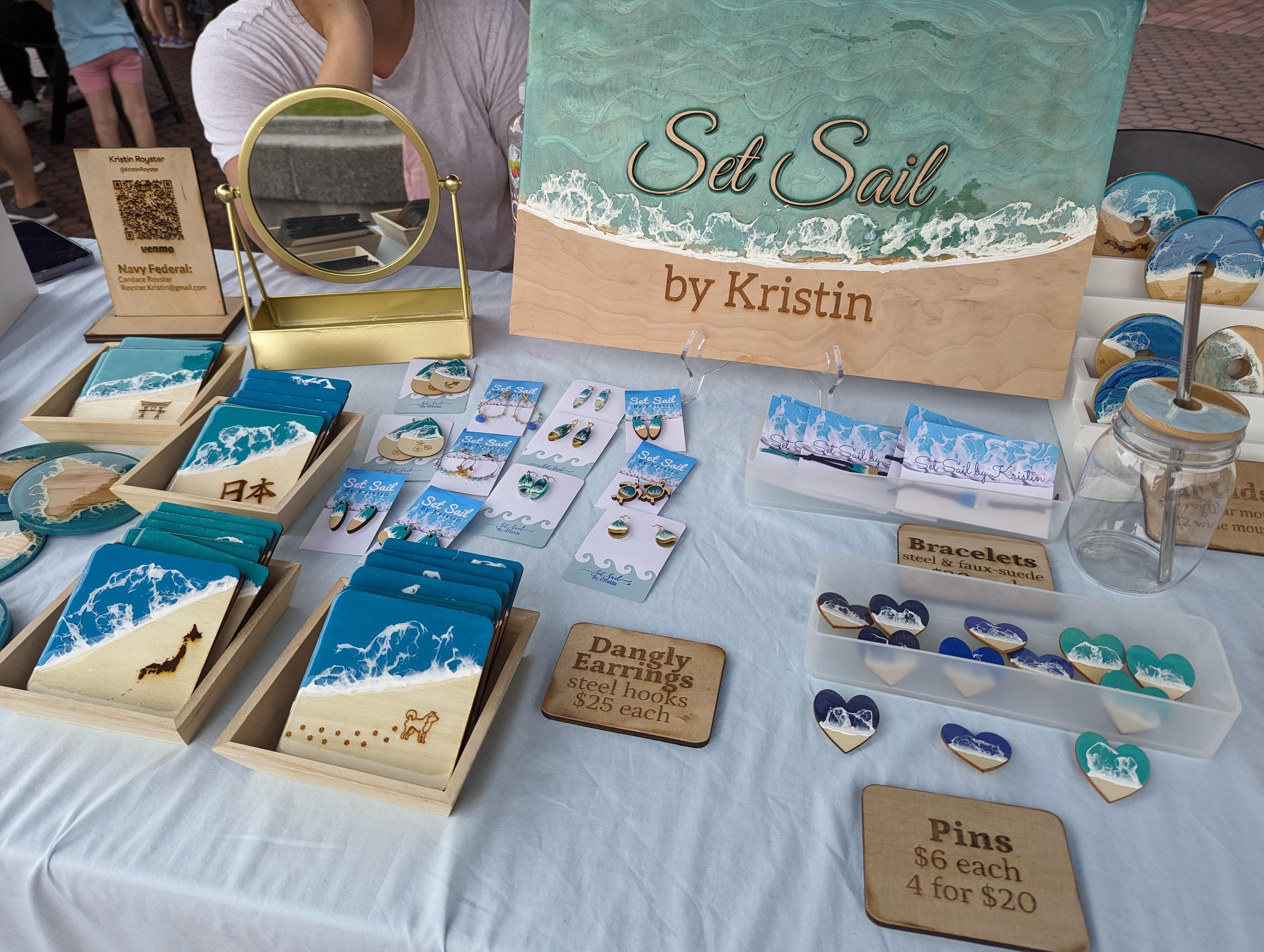 Table displays jewelry and accessories with a sign reading 'Sea Sail by Kristin' and a small wooden sign about pins.