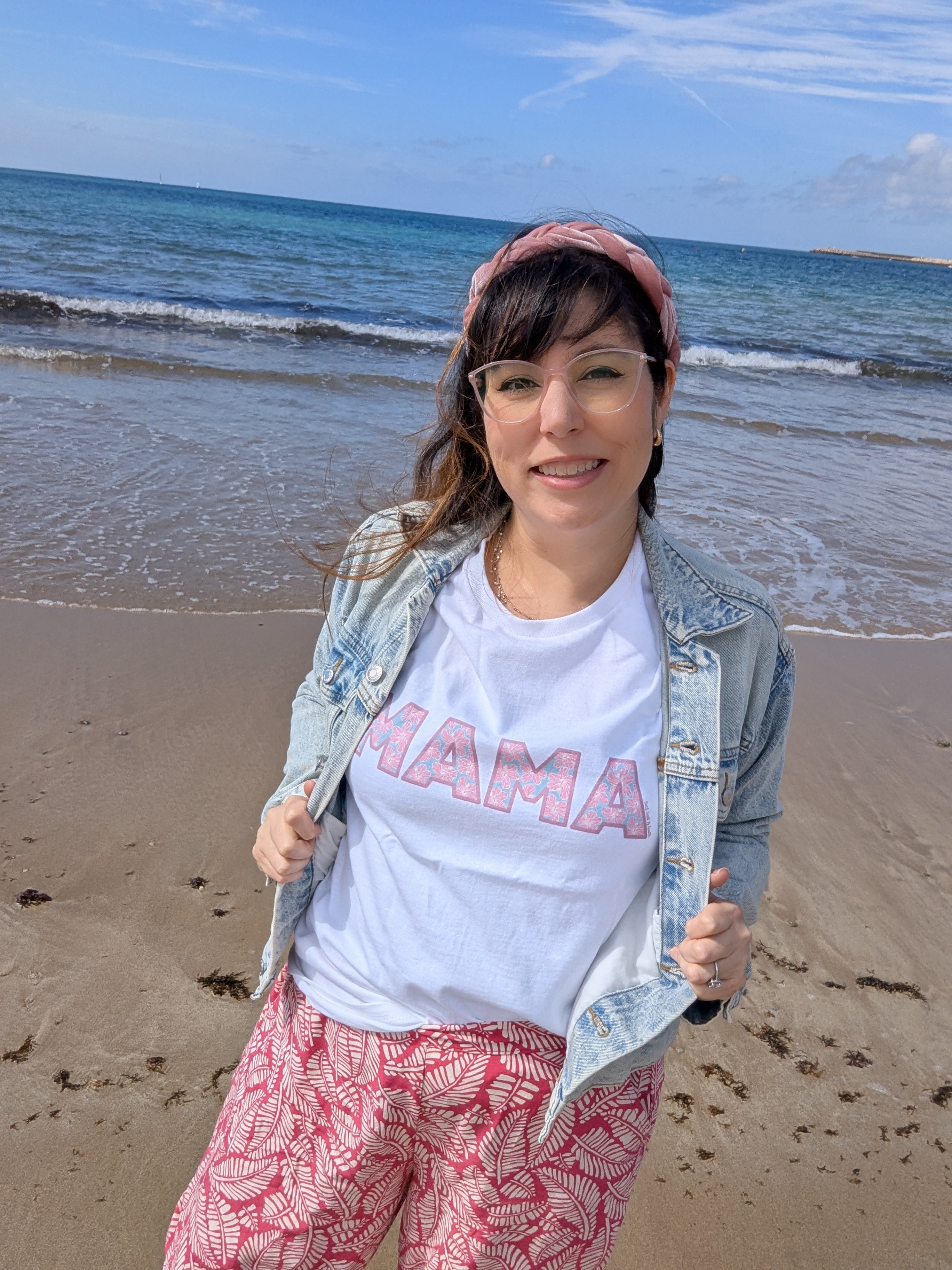 Woman smiling on beach, wearing a white T-shirt with pink text, denim jacket, and patterned pants, with ocean and sky in background.