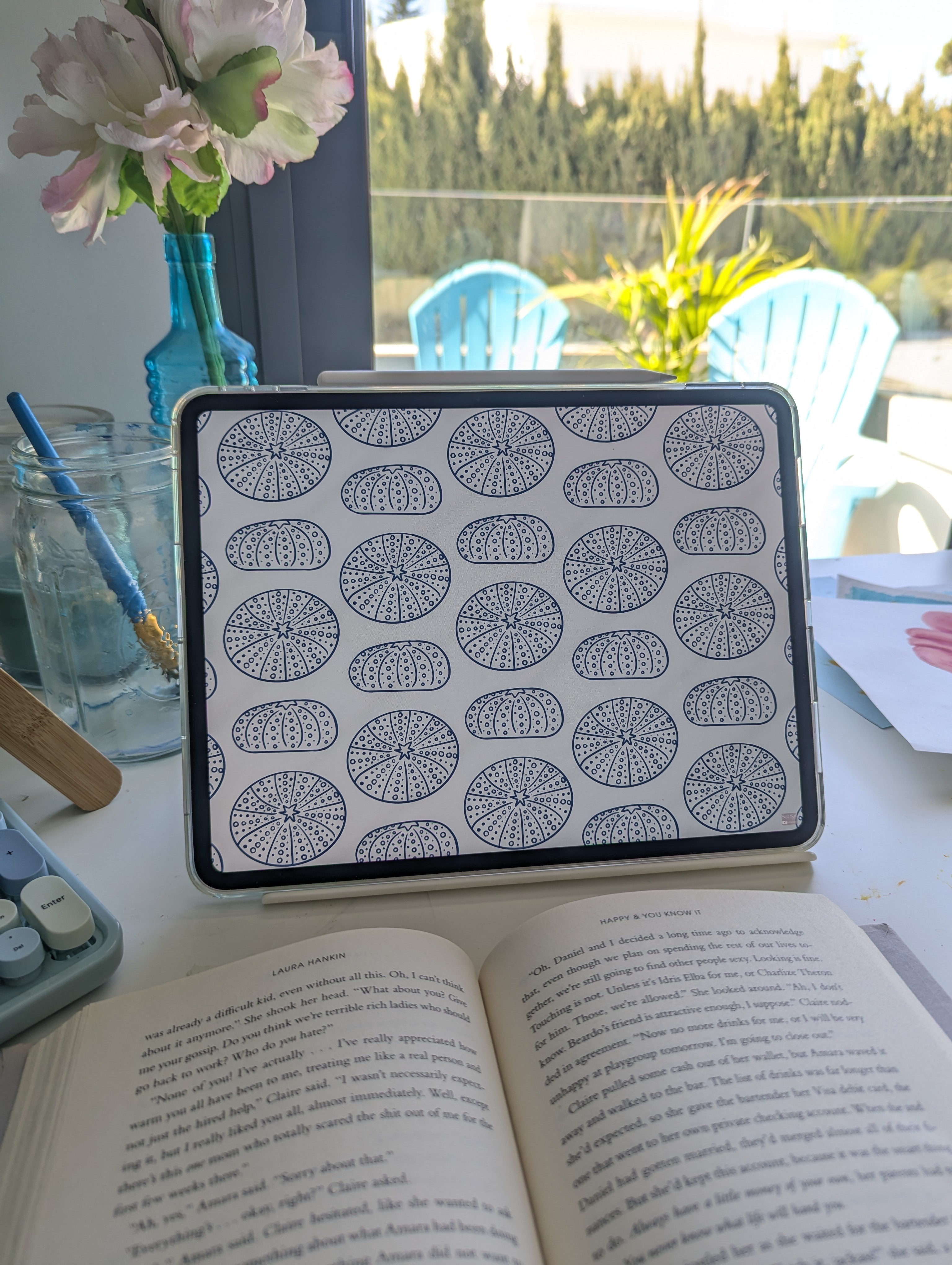 Open book on table with patterned tablet or book cover behind it, window with outdoor view in background.