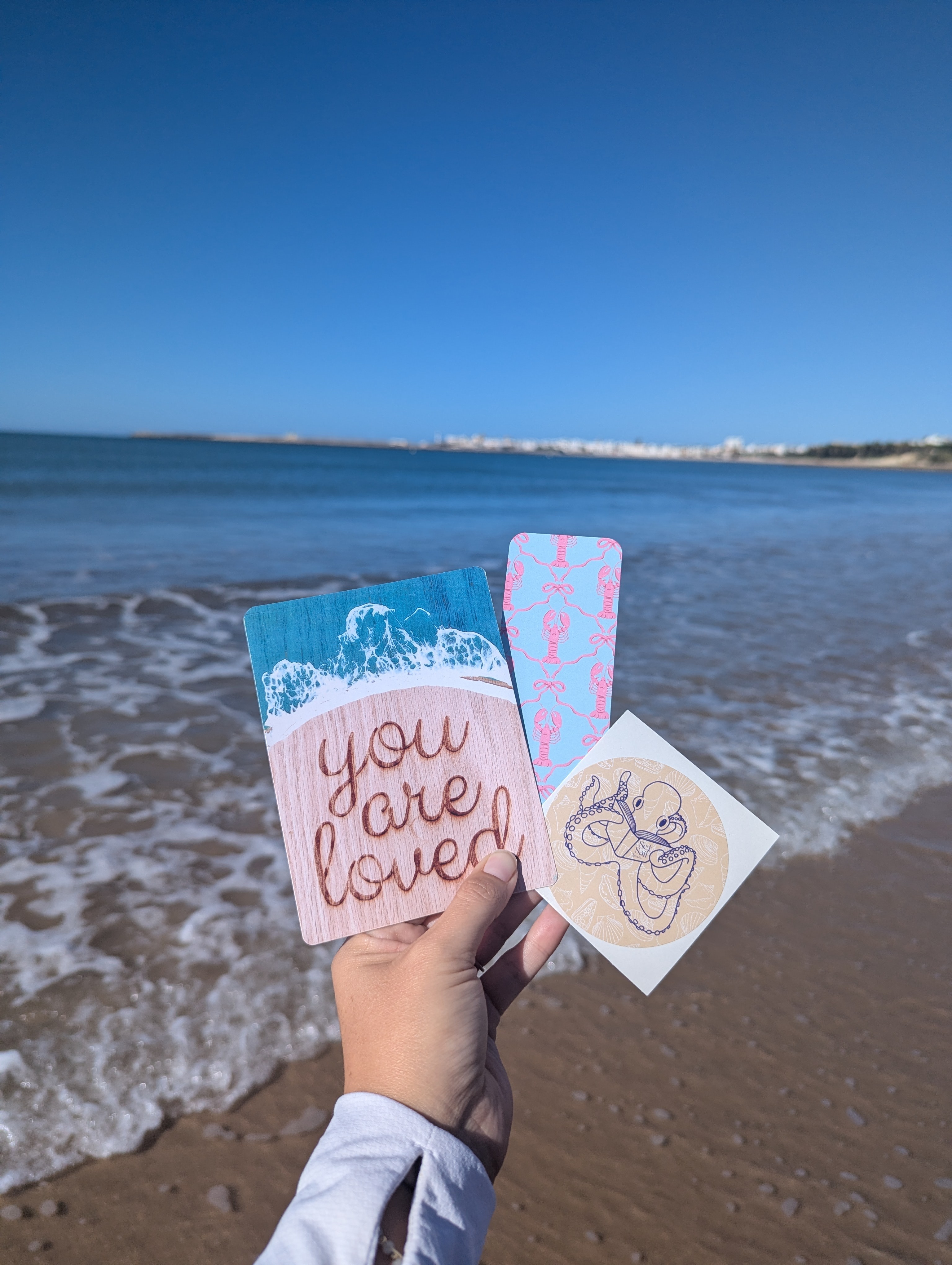 Person holding three small cards with beach and ocean in background, one card says 'you are loved', others have designs.