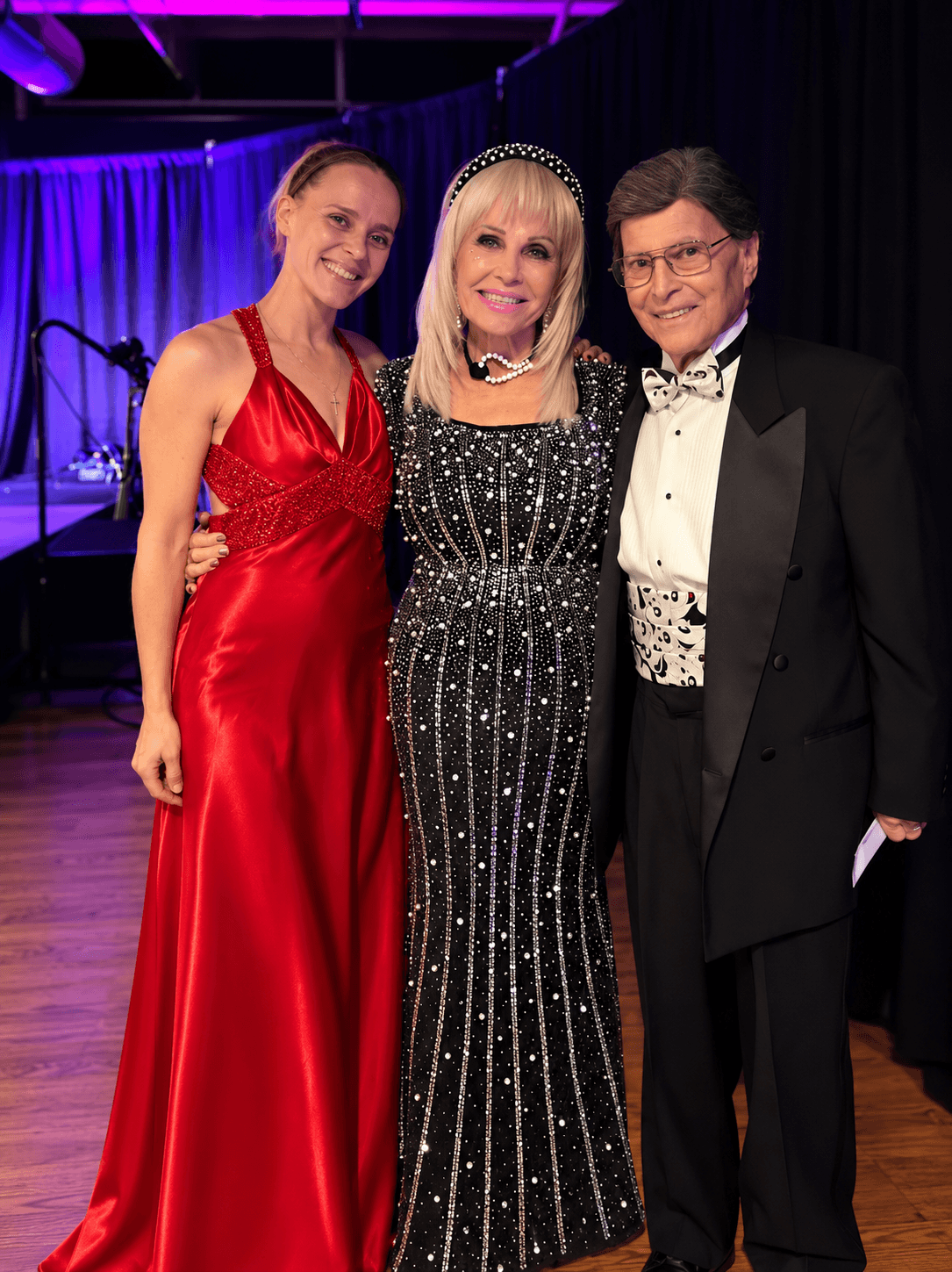 Three people in formal attire standing on stage, smiling, with a dark curtain background.