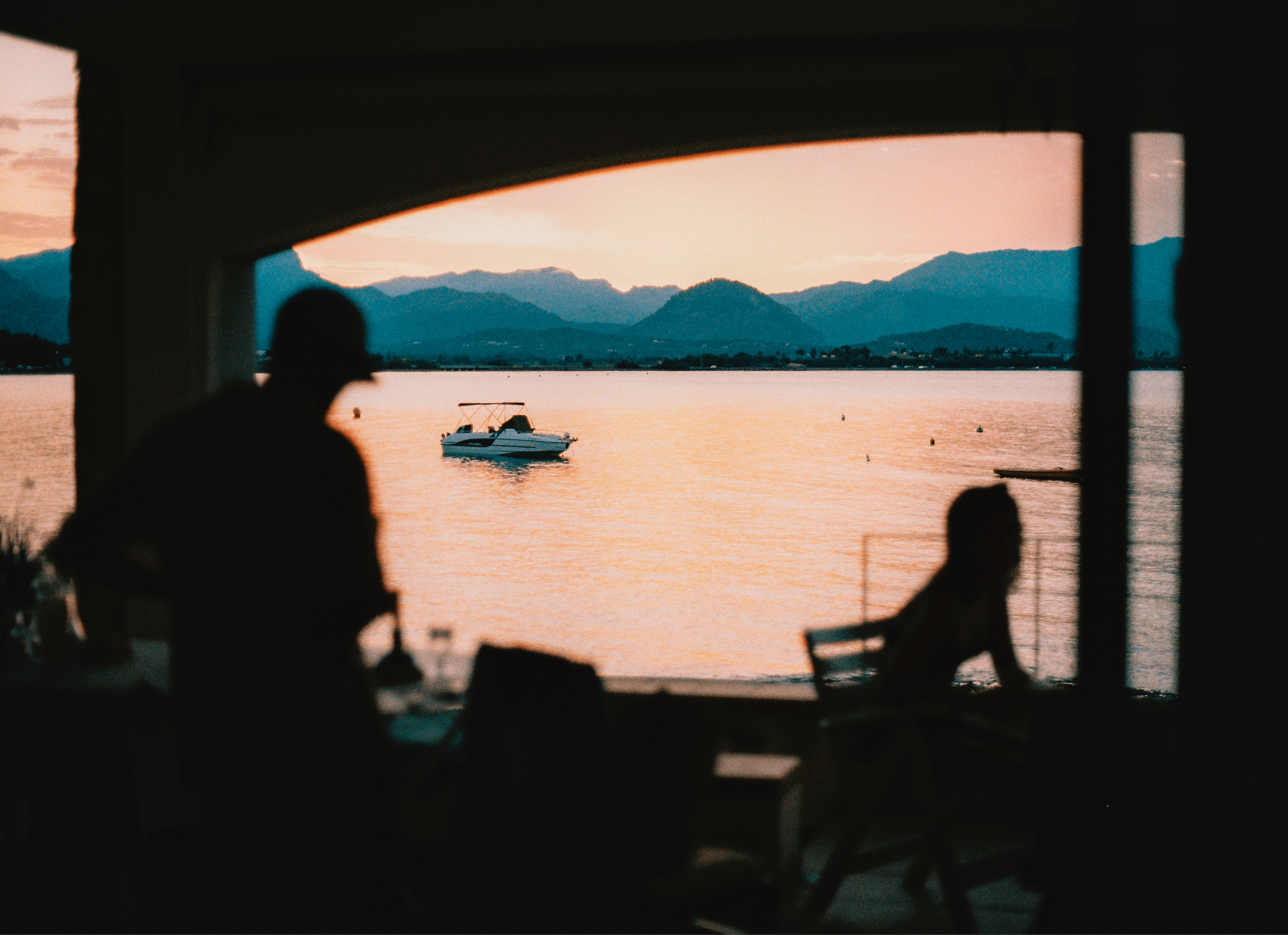 Silhouettes of two people near a window overlooking a lake with a boat, mountains, and sunset sky in the background.