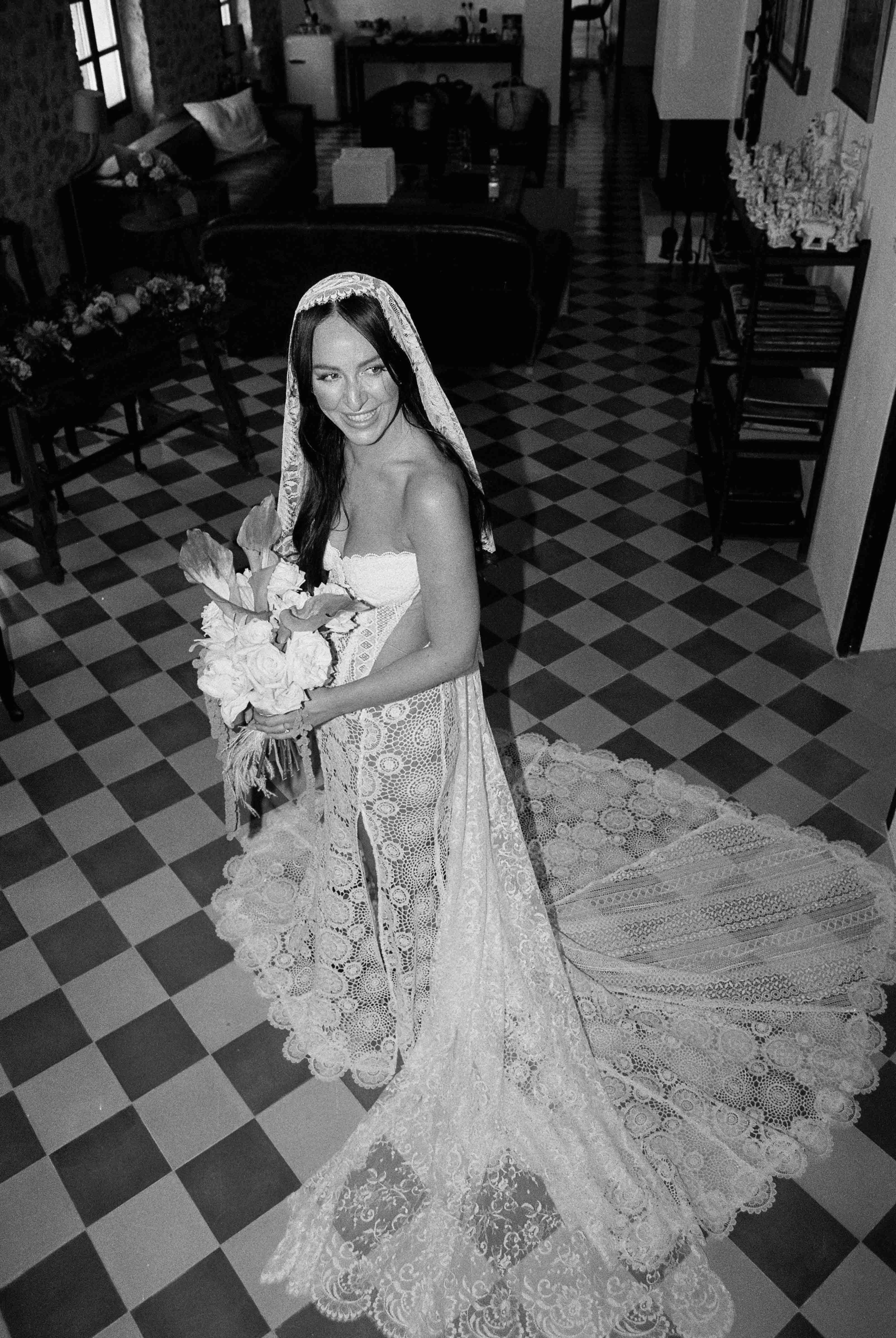 Bride in wedding dress holding bouquet, standing in a checkered floor room with chairs and furniture, smiling at camera.