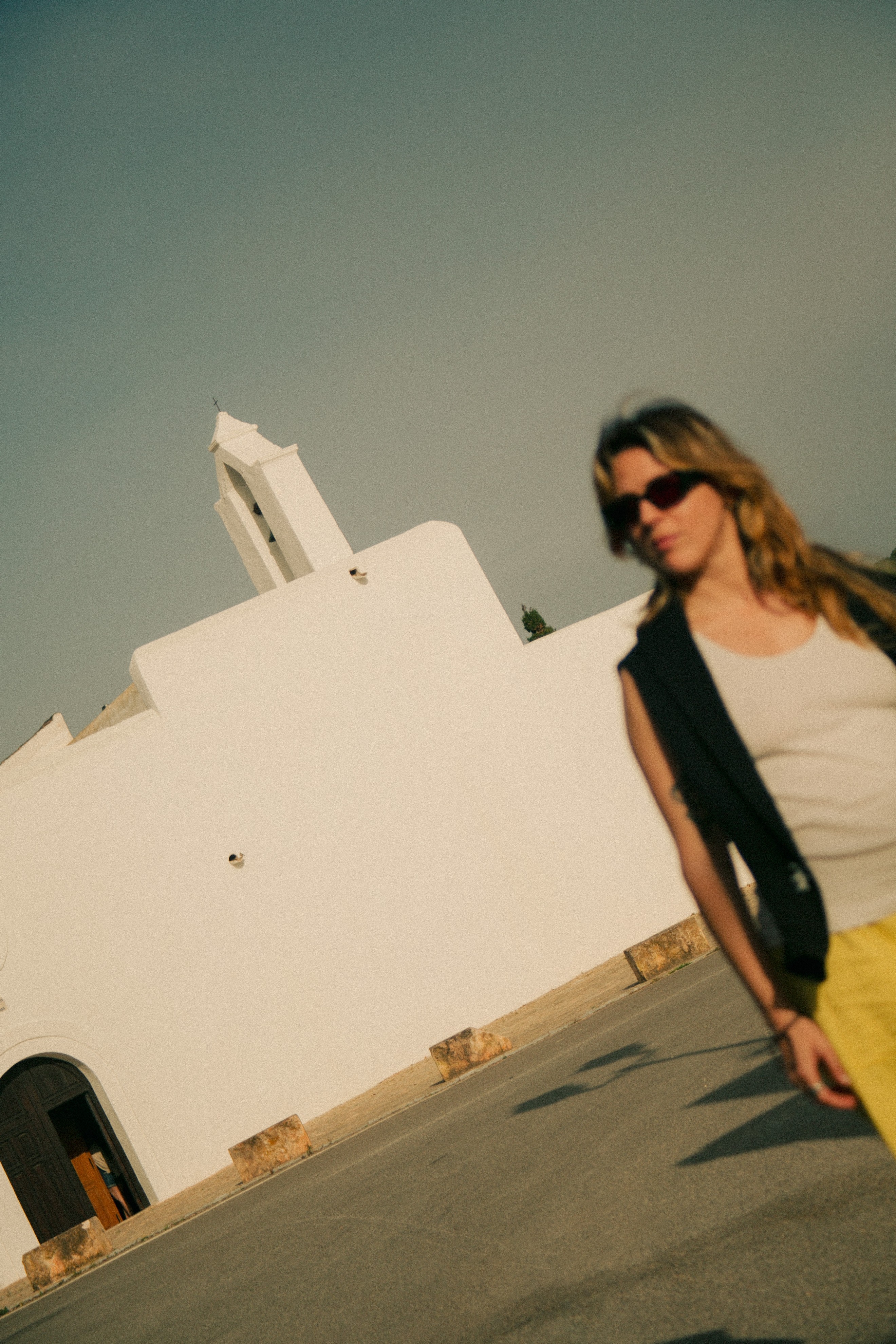 Woman with sunglasses and a vest standing outdoors near a white building under a cloudy sky.