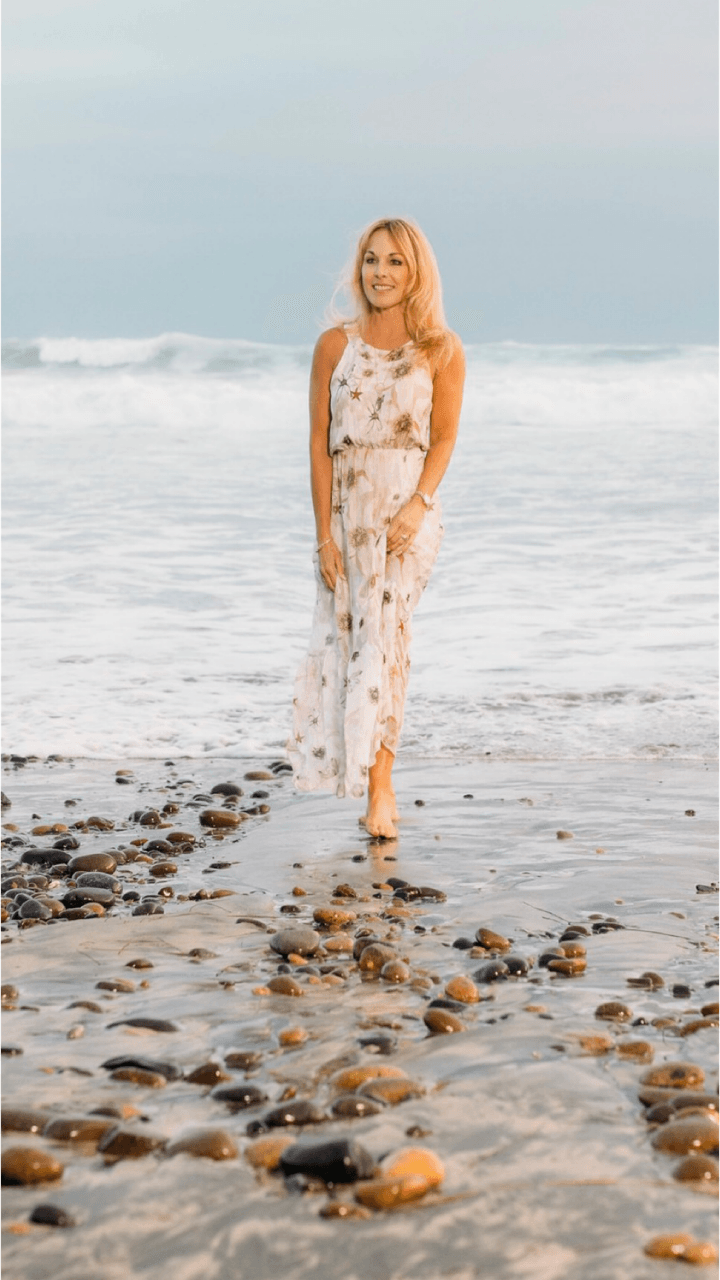 Woman in a white floral dress walking on a rocky beach with waves in the background.