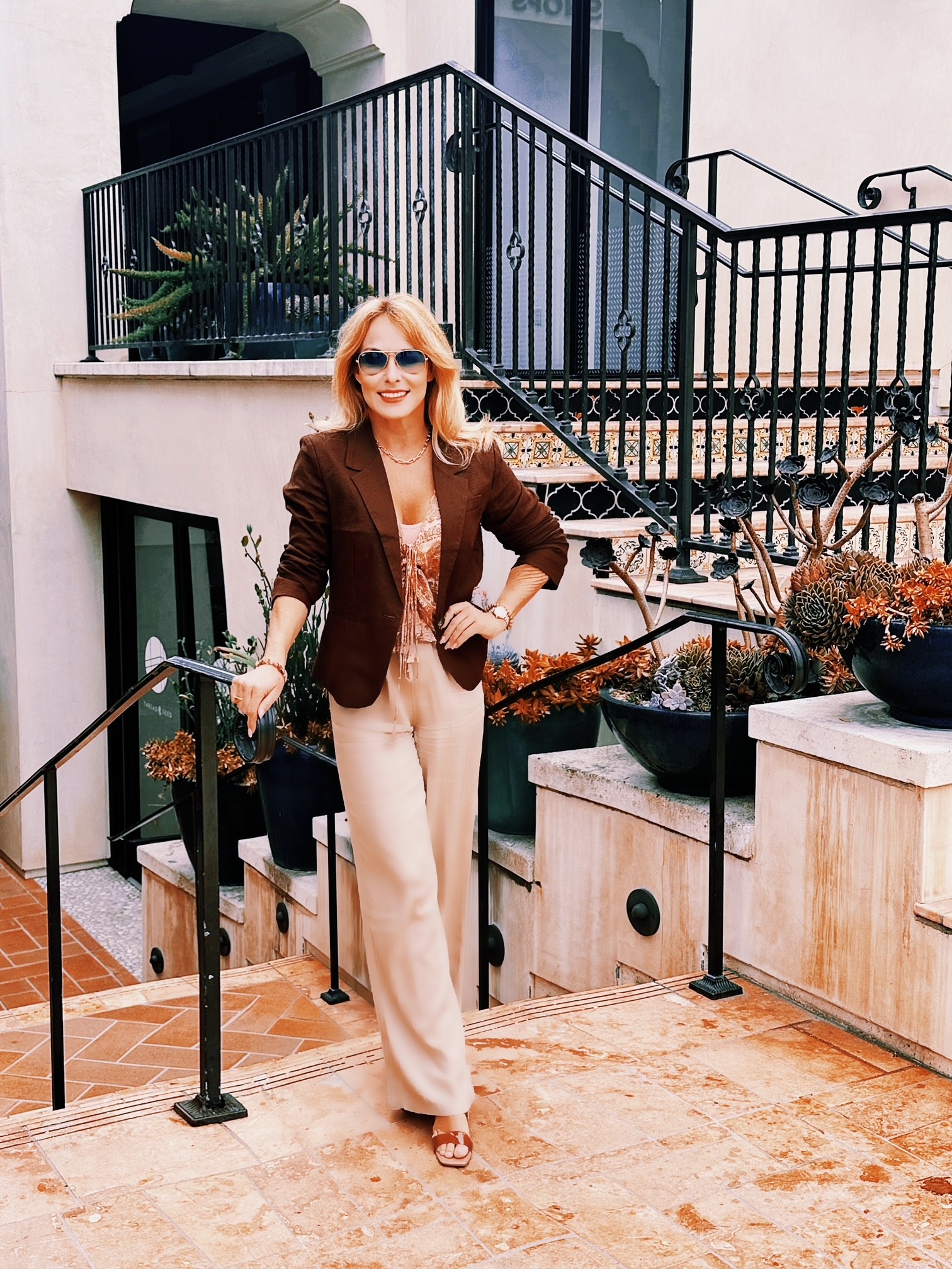 Woman with sunglasses and blonde hair in a brown blazer and beige pants standing outdoors near stairs and railing.