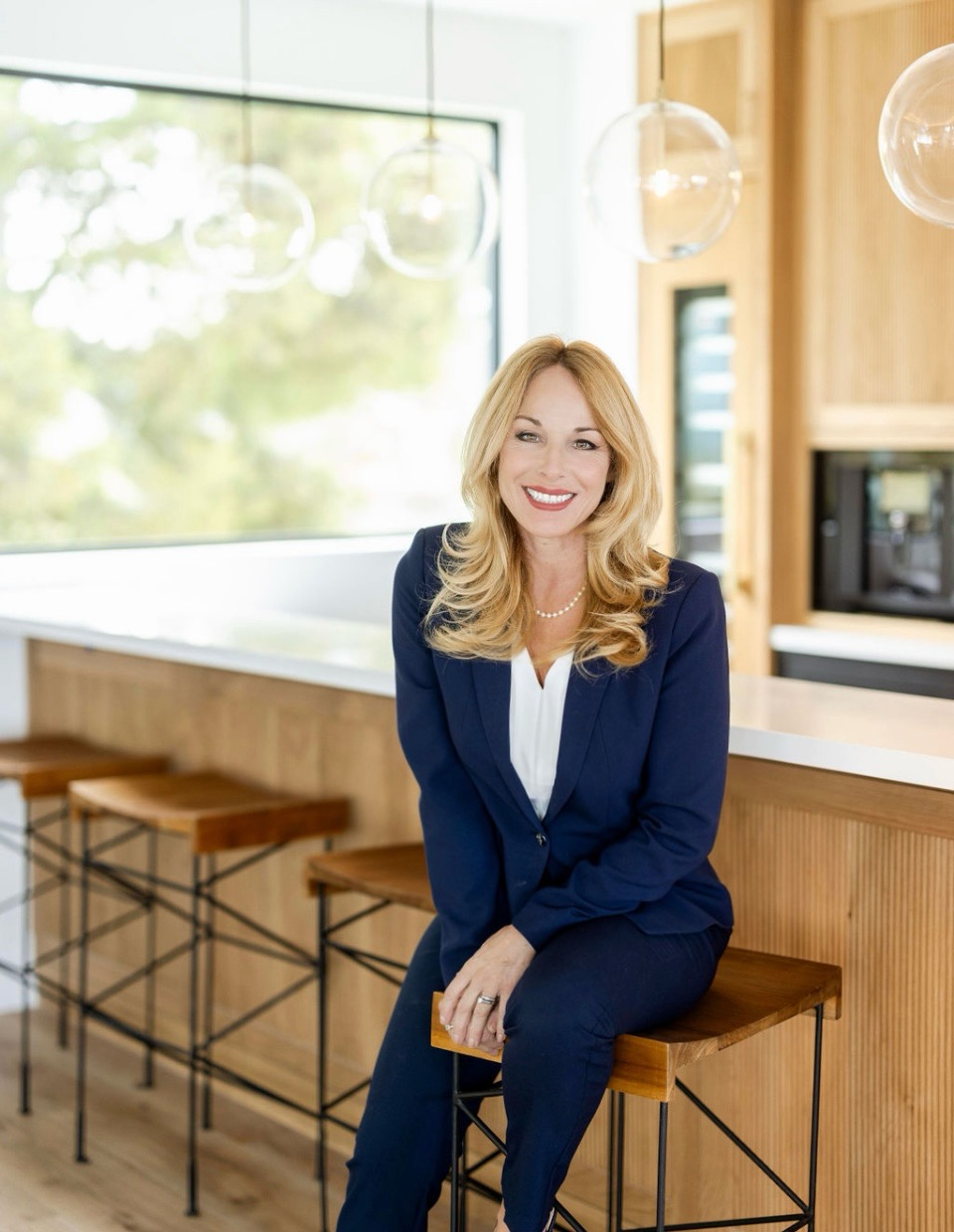 Woman in navy suit sitting on a wooden barstool in a modern kitchen, smiling, with large window and hanging lights behind her.