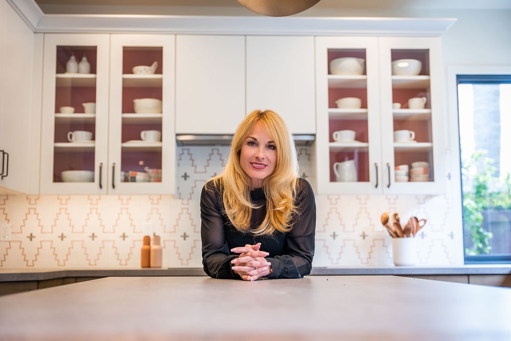 Woman with blonde hair smiling, leaning on kitchen counter with white cabinets and open shelves behind her.