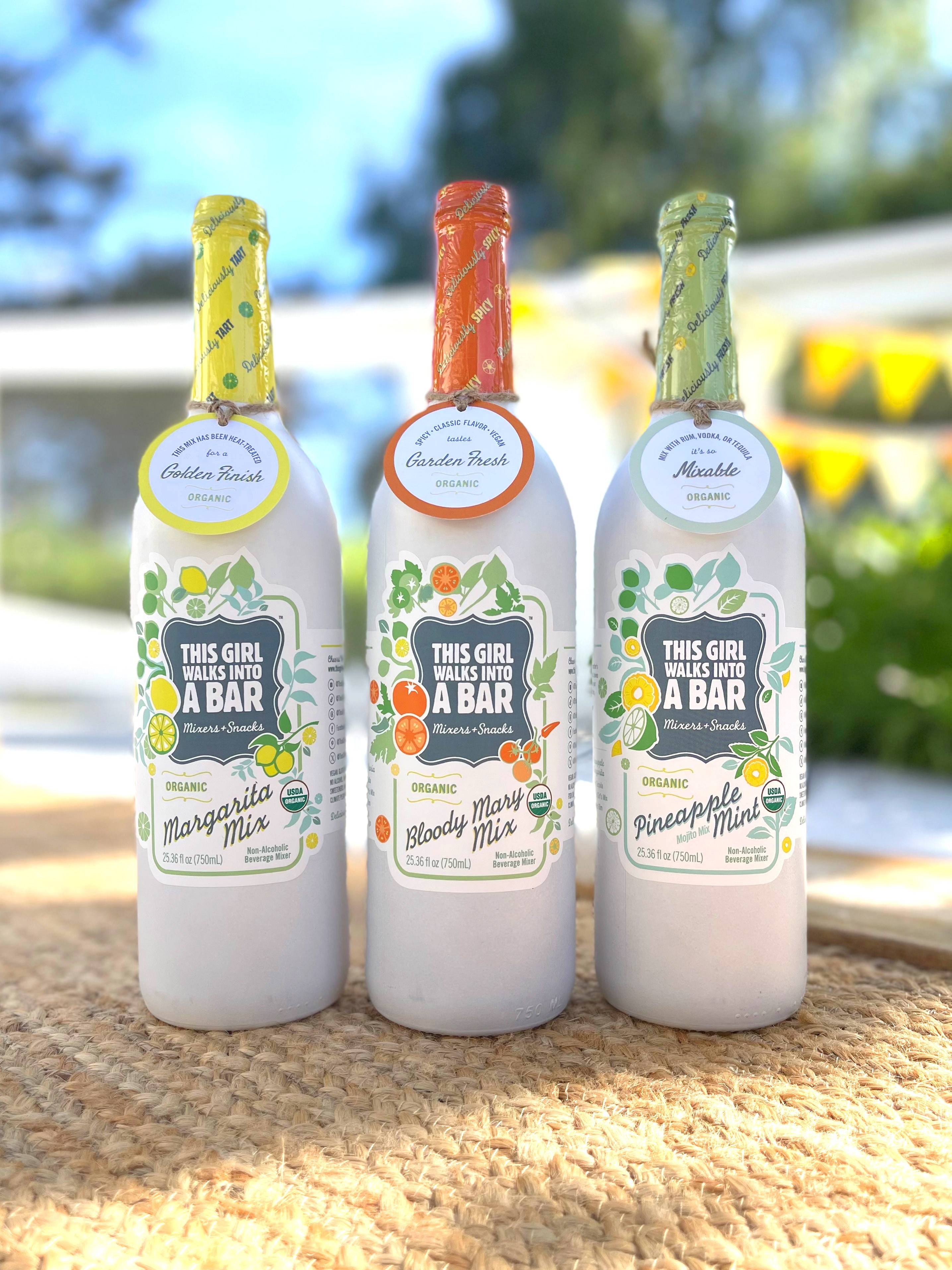 Three bottles with colorful caps and labels, placed outdoors on a textured surface, with trees and sky in the background.