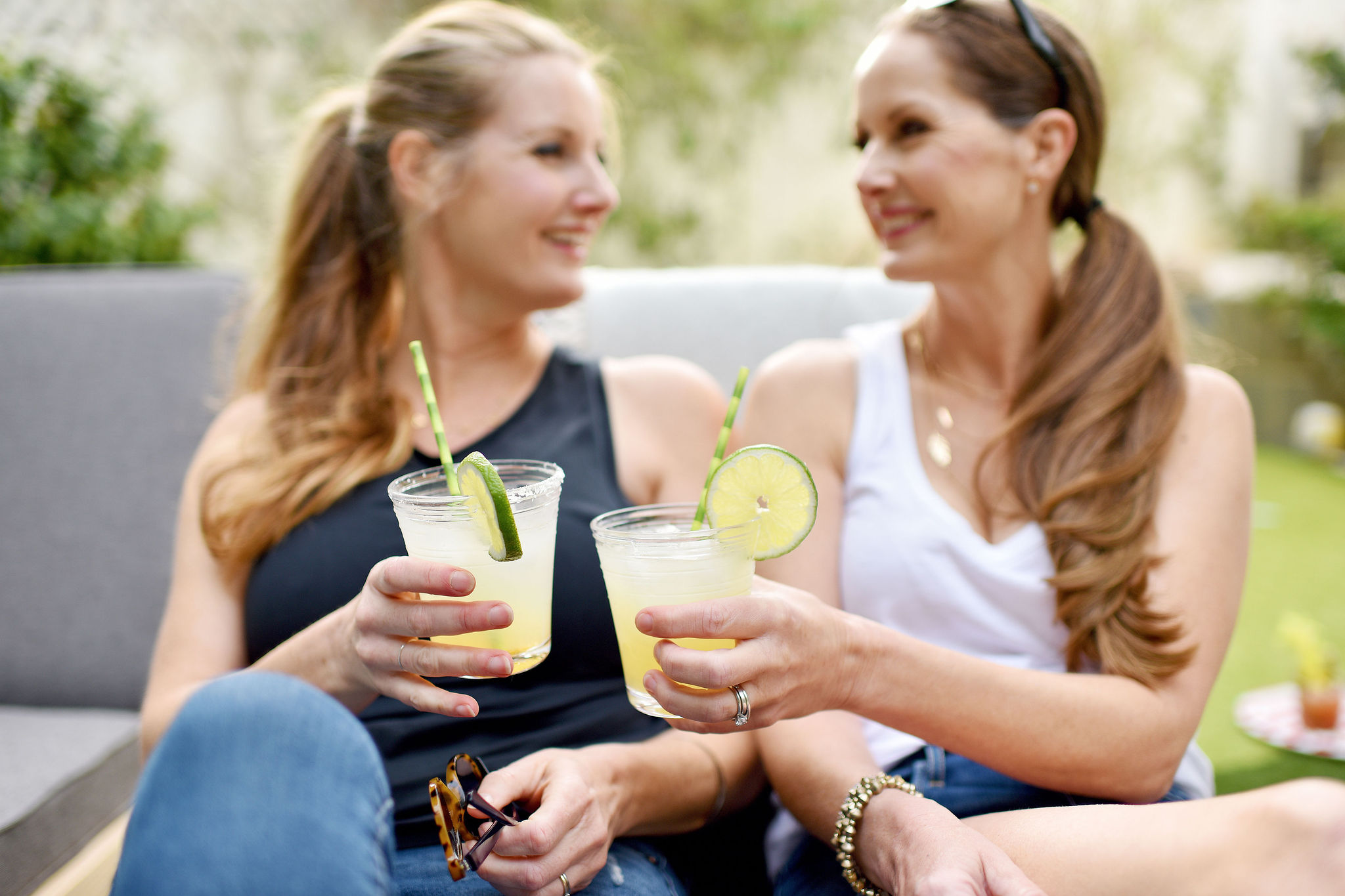 Two women sitting outdoors, smiling, holding drinks with lime slices, engaging in conversation, with greenery in the background.