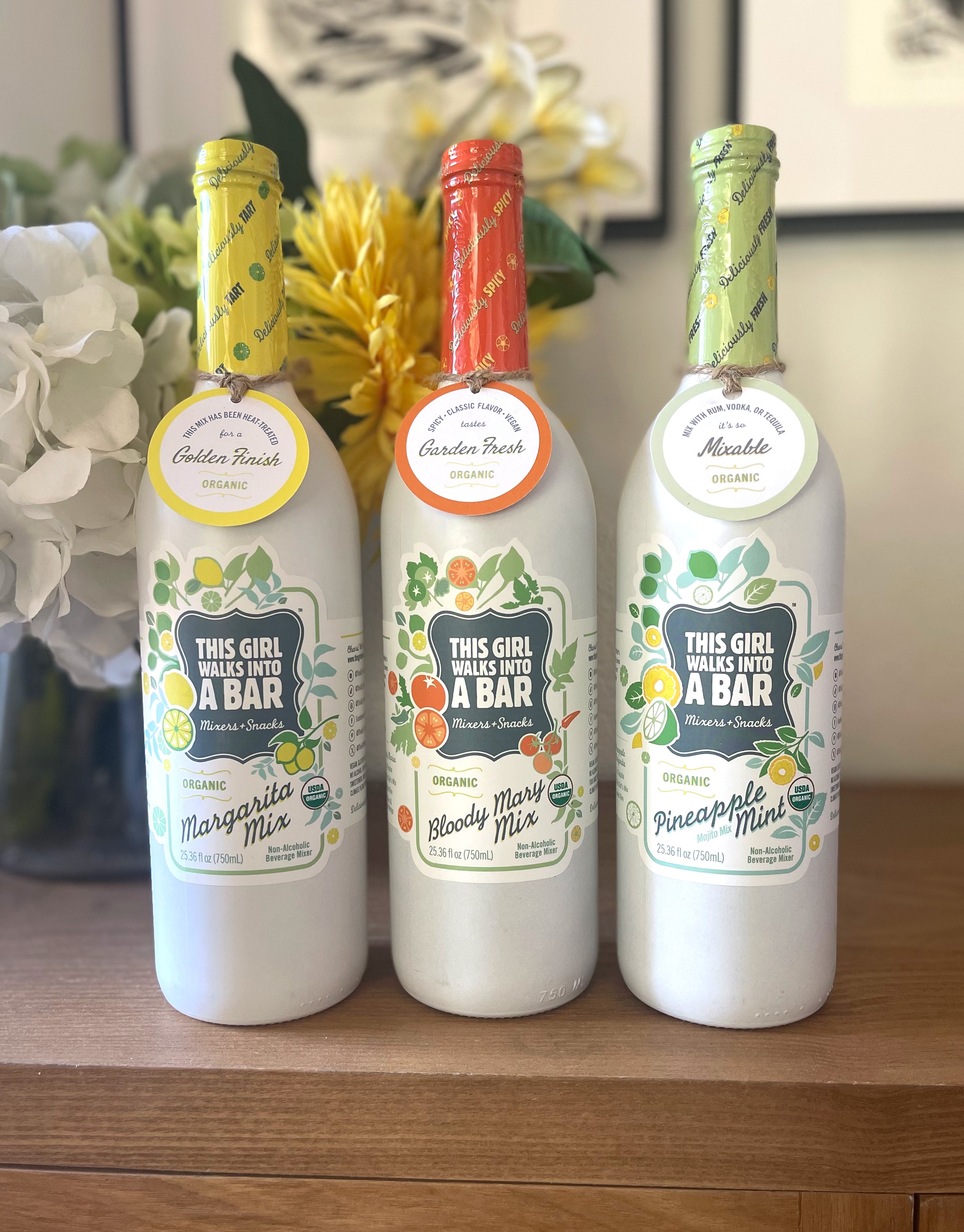 Three bottles with colorful caps and labels, placed on a wooden surface with flowers in the background.
