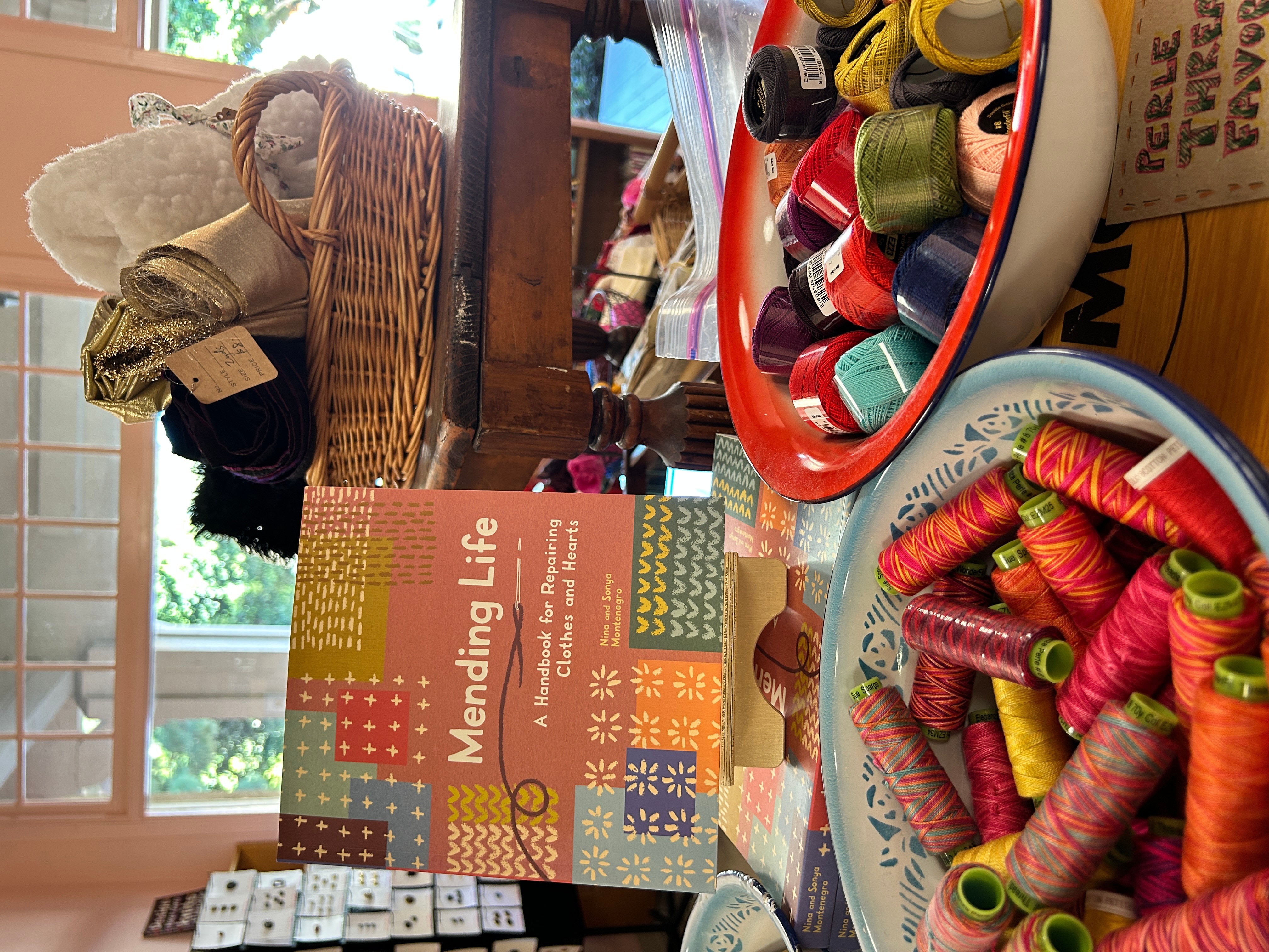 Display of sewing supplies including spools of thread, a basket, and a book titled 'Mending Life' on a table.