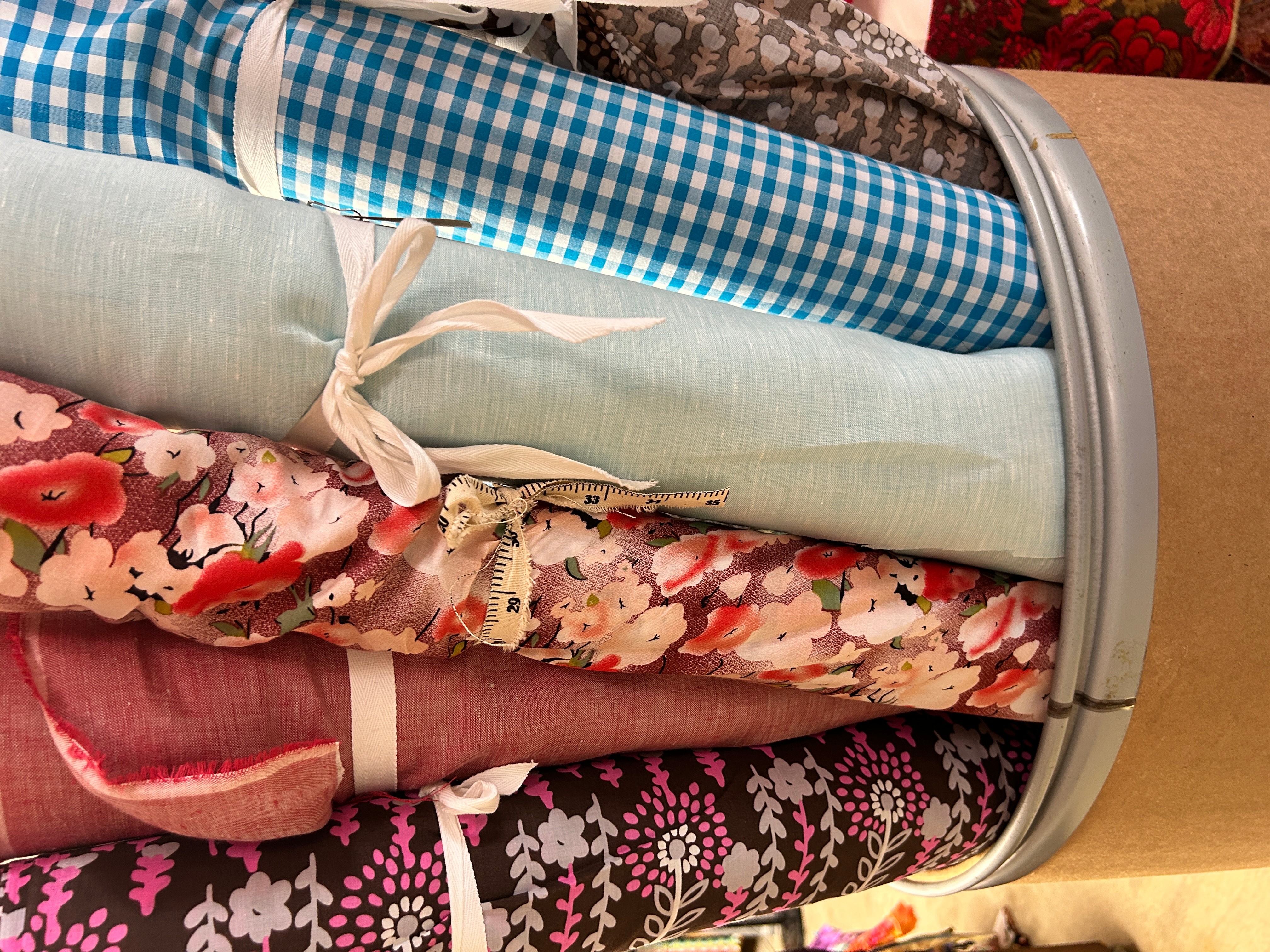 Stacked fabric rolls with various patterns and colors, including checkered, floral, and solid fabrics, on a metallic rack.