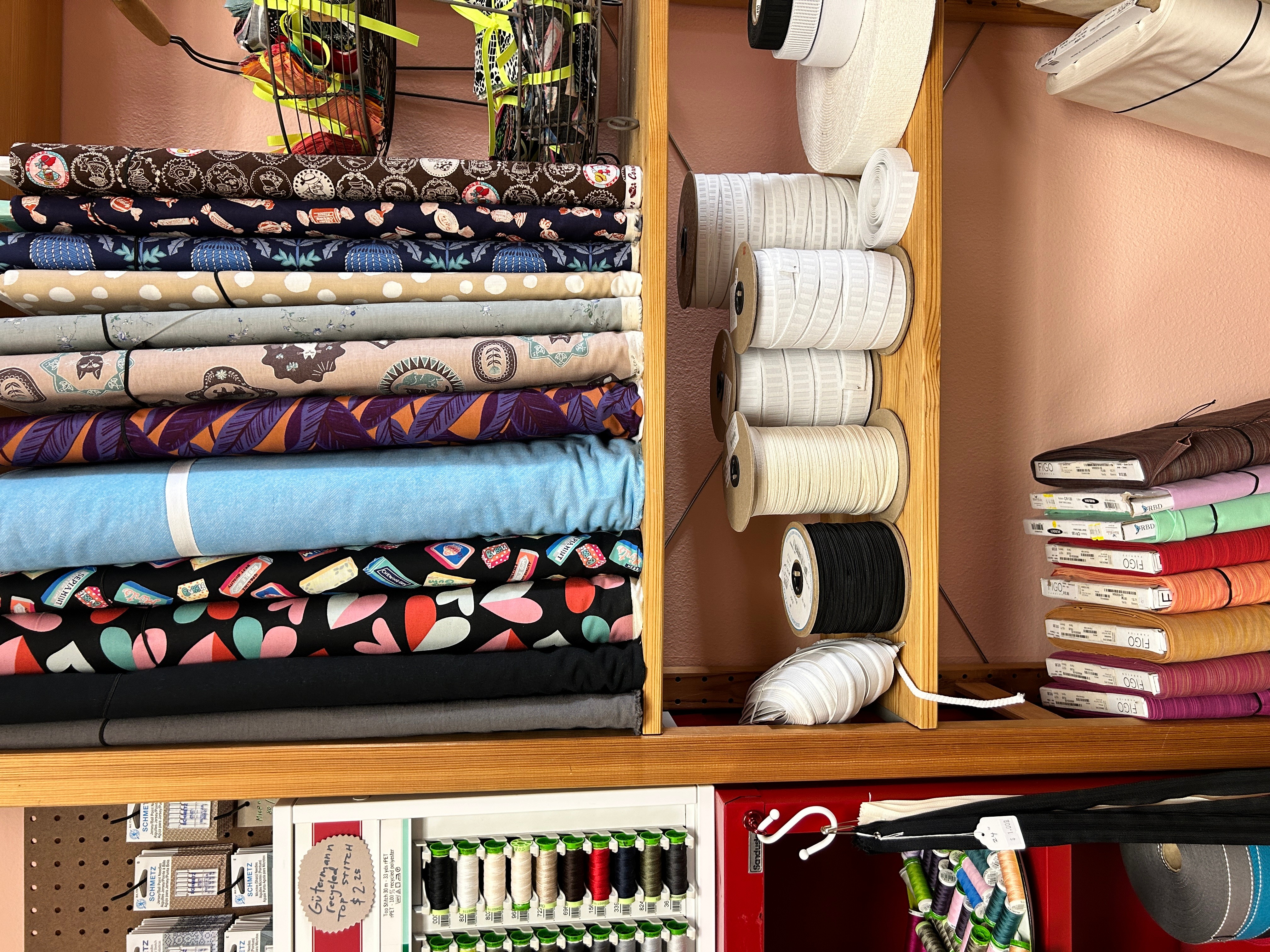 Shelves with fabric rolls, sewing threads, and stacked books or notebooks, in a sewing or craft room.