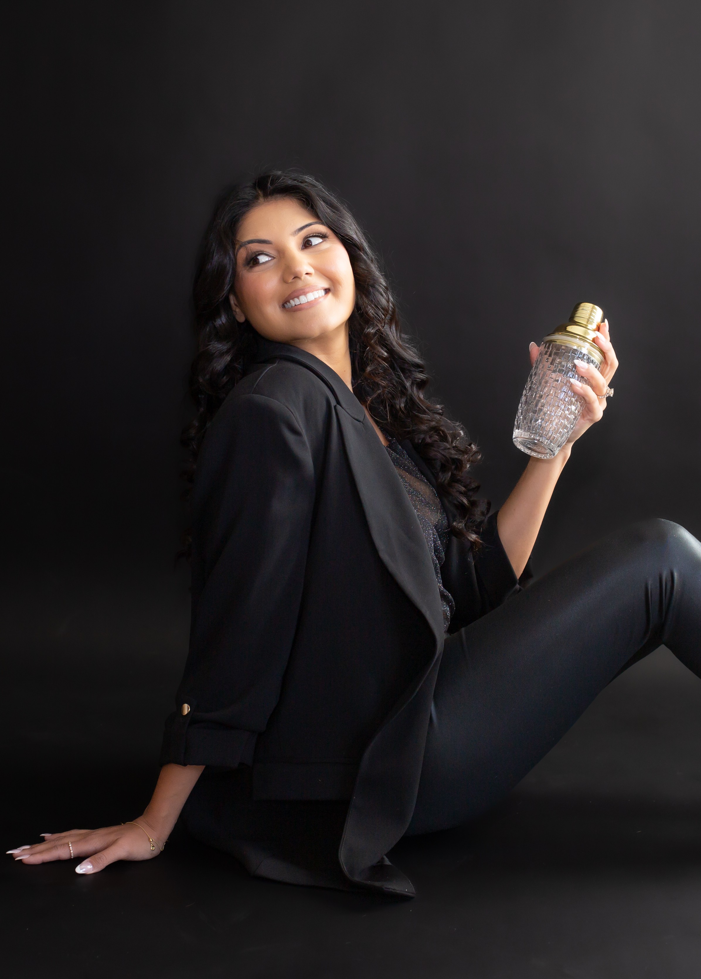 Smiling woman with long dark hair sitting on the floor holding a glittery water bottle, against a dark background.