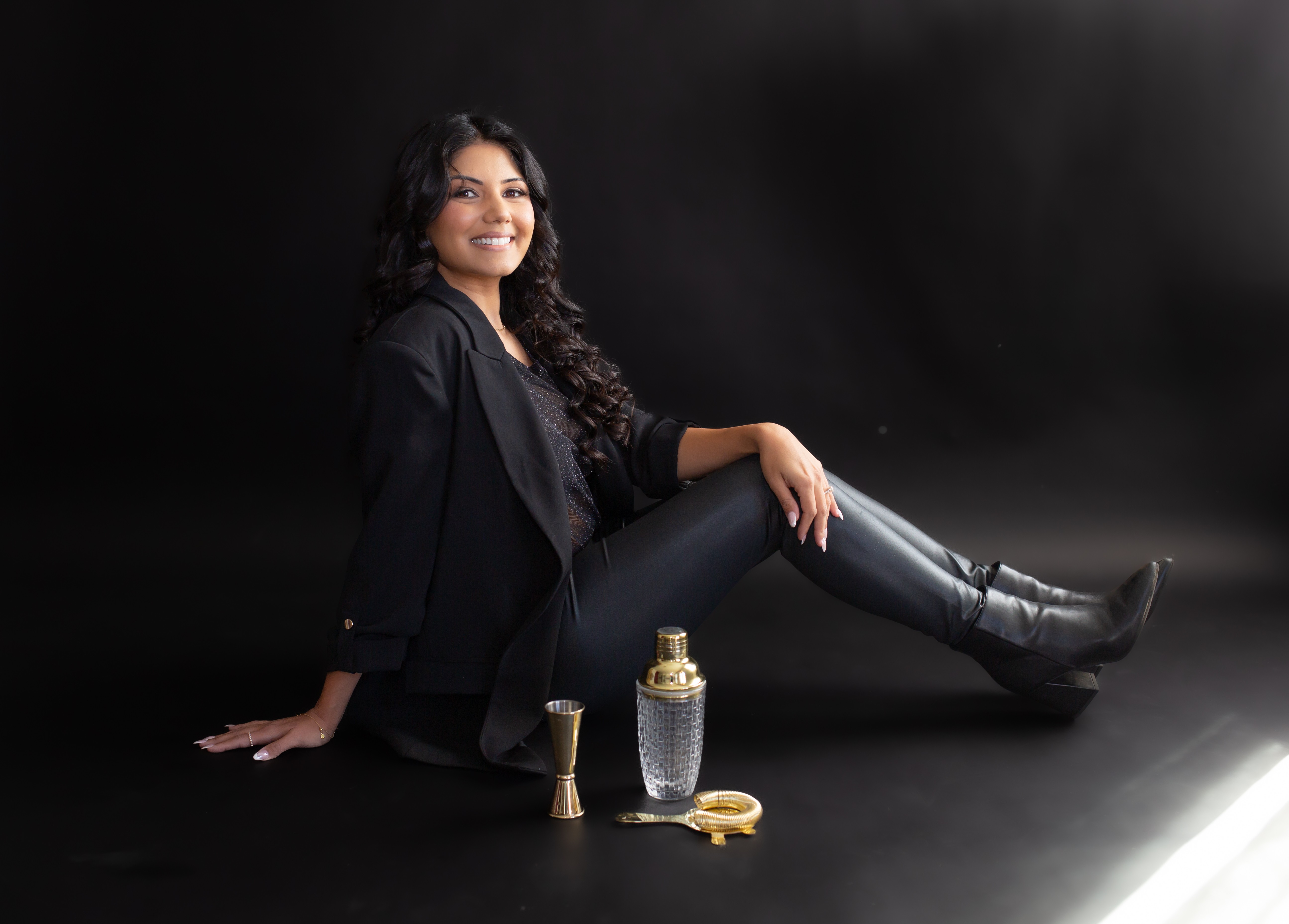 Woman with long curly hair sitting on the floor, smiling, with perfume bottles and jewelry in front, against a black background.