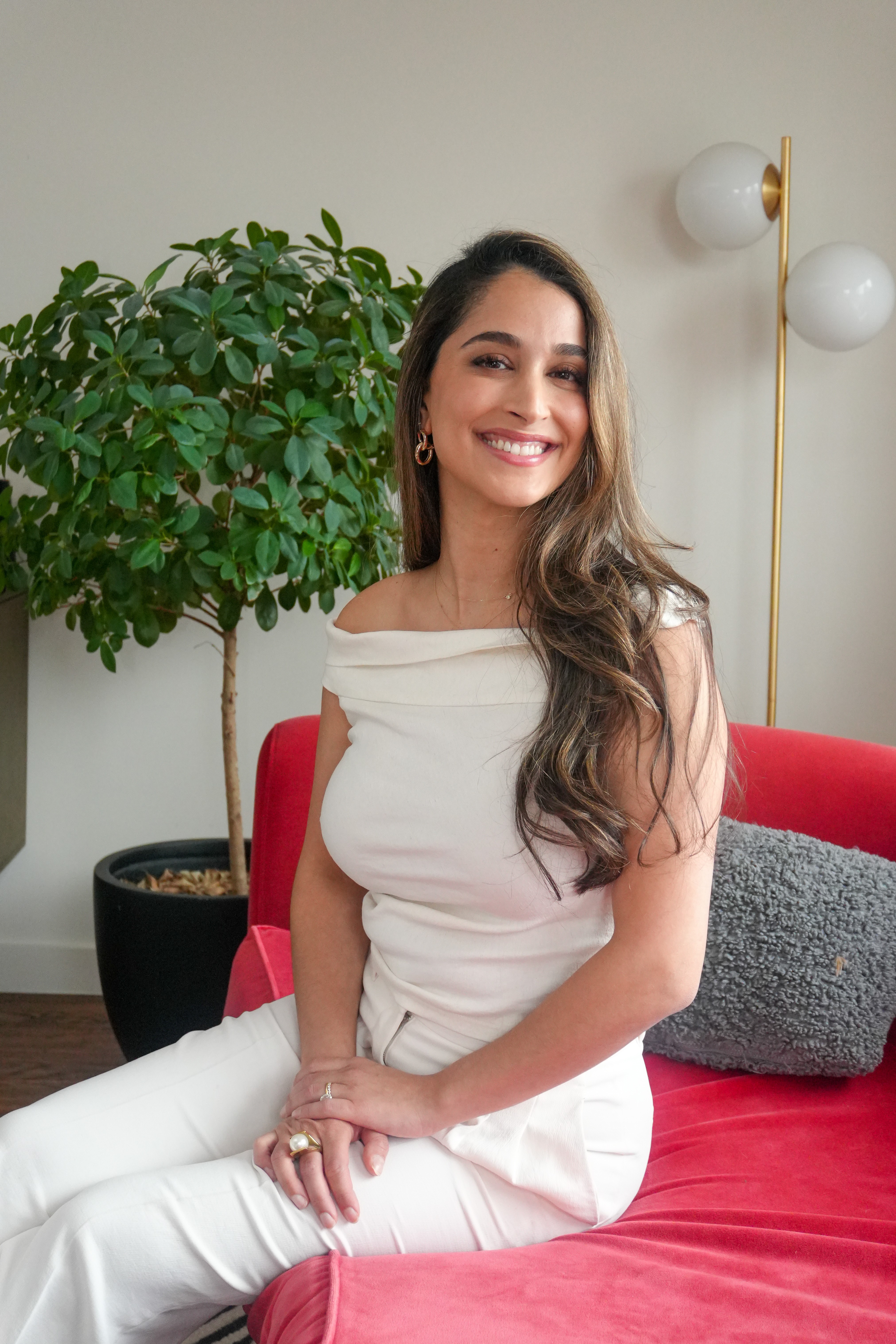 Smiling woman with long wavy hair sitting on a red couch in a room with a large green plant and a modern wall lamp.