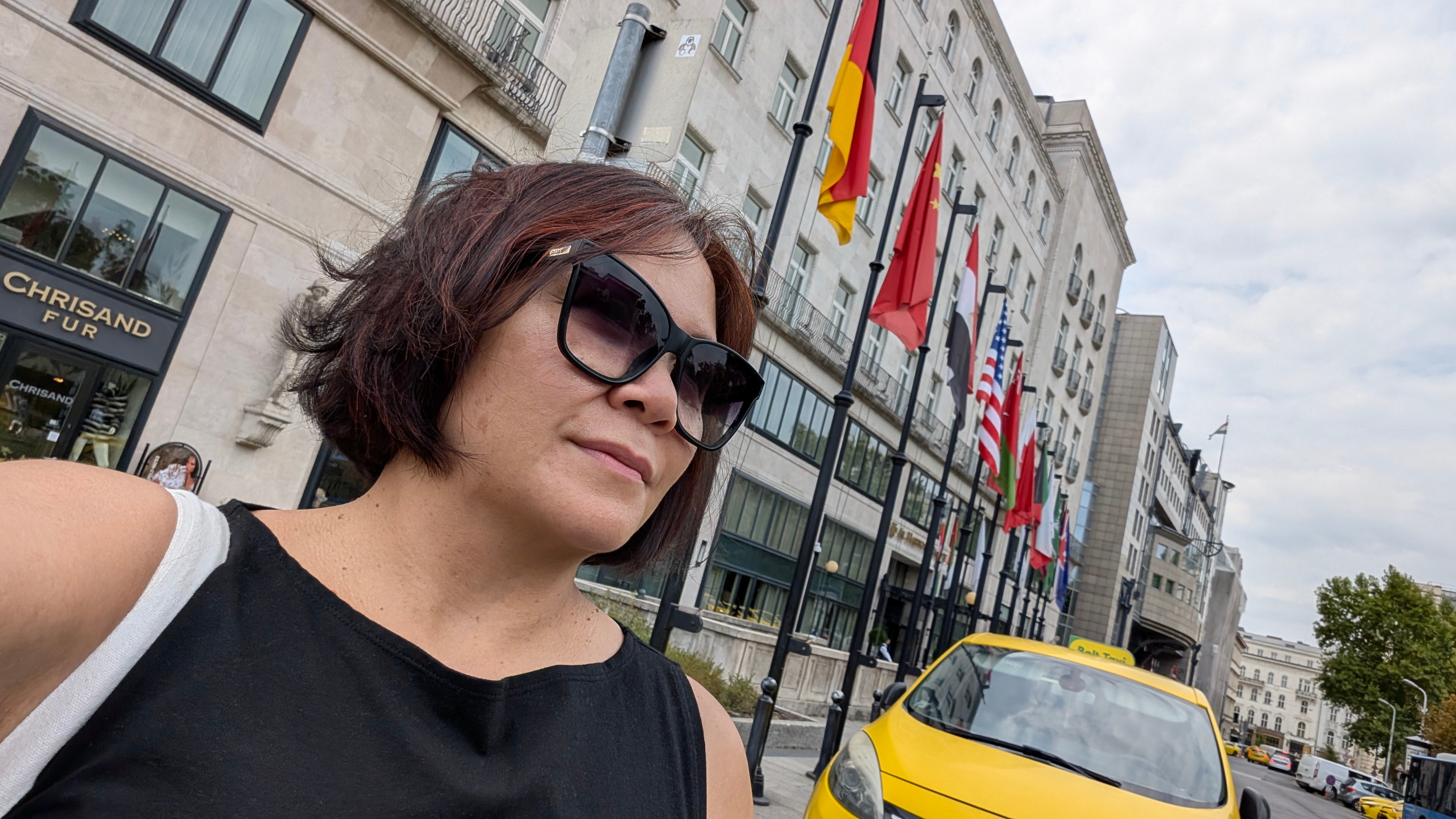 Woman with short hair and sunglasses taking a selfie on a city street with flags and buildings in the background.