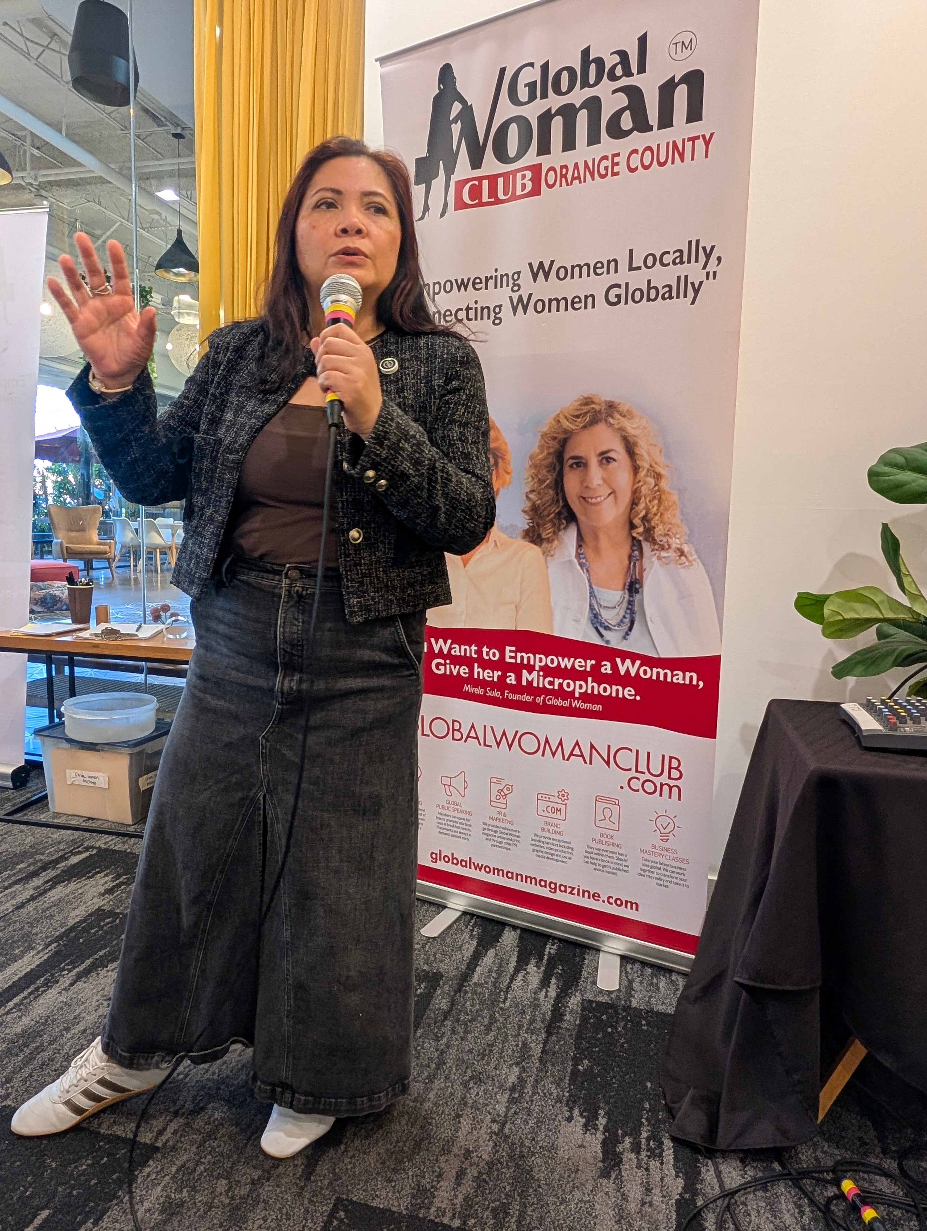 Woman speaking into microphone at event, standing next to a banner for Global Woman Club Orange County, with a woman on the banner.