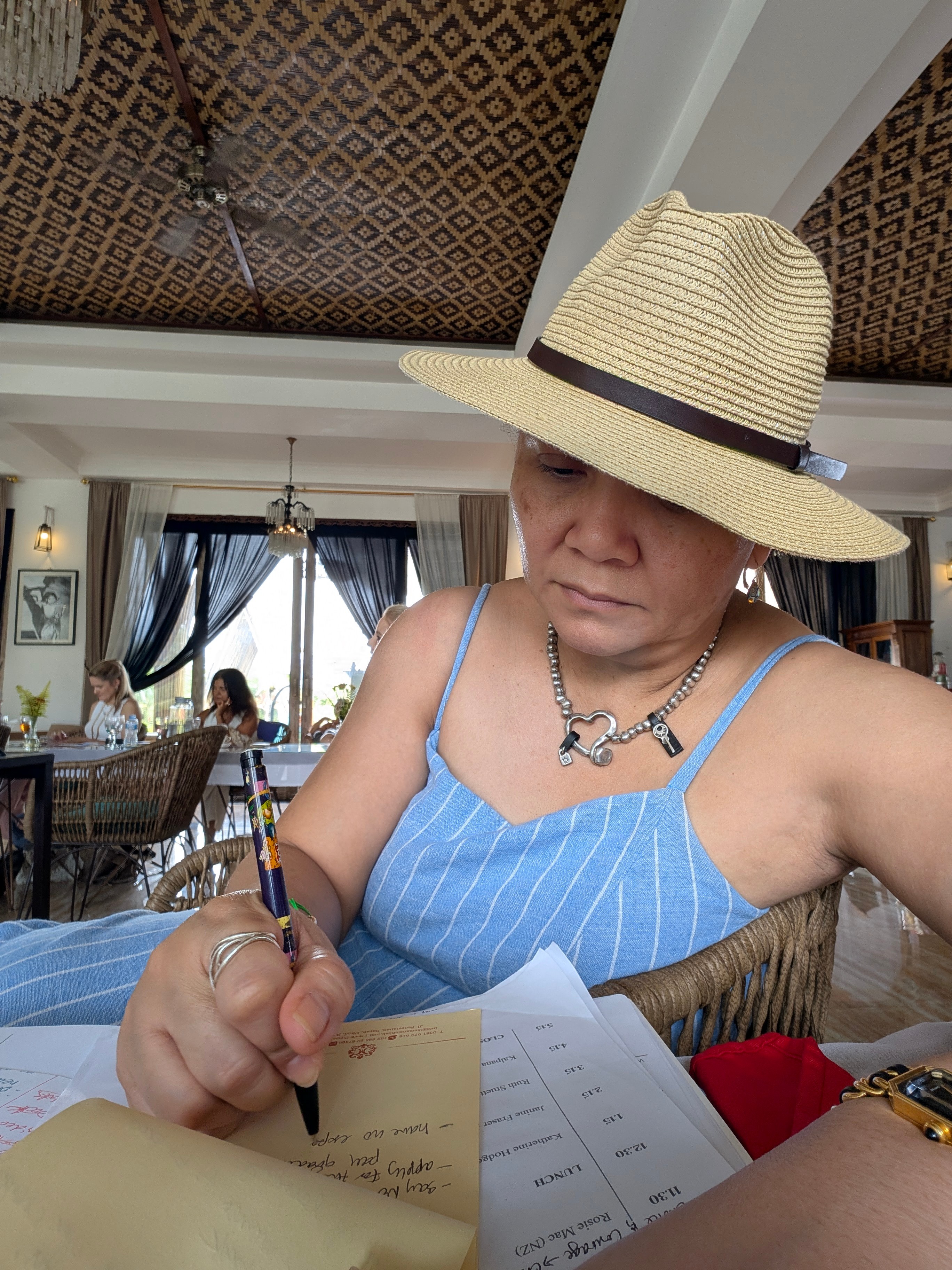 Woman wearing a straw hat and blue striped dress writing on paper in a well-lit room with curtains and furniture