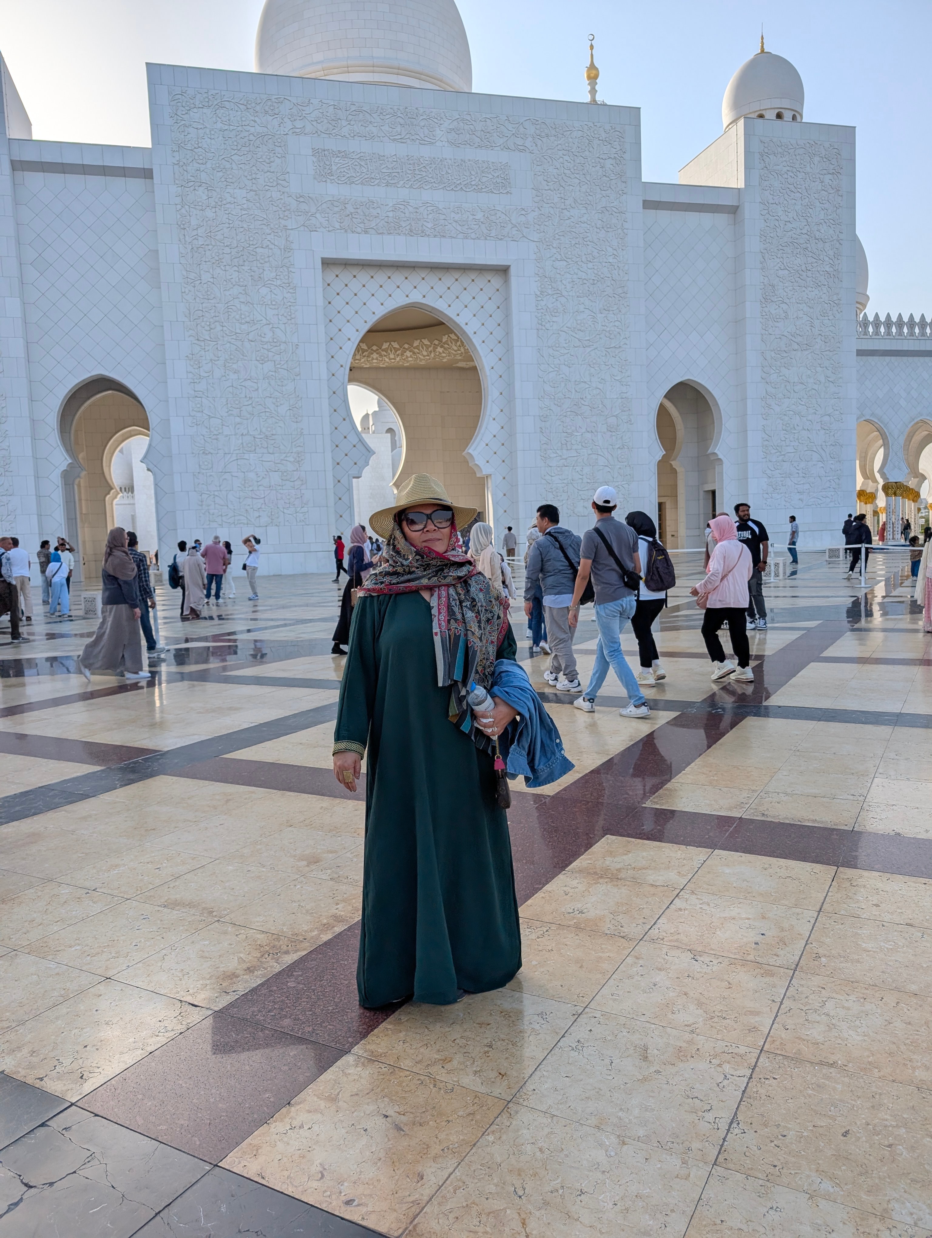 Woman in a long dress and headscarf standing in front of a large white building with arches, surrounded by people.