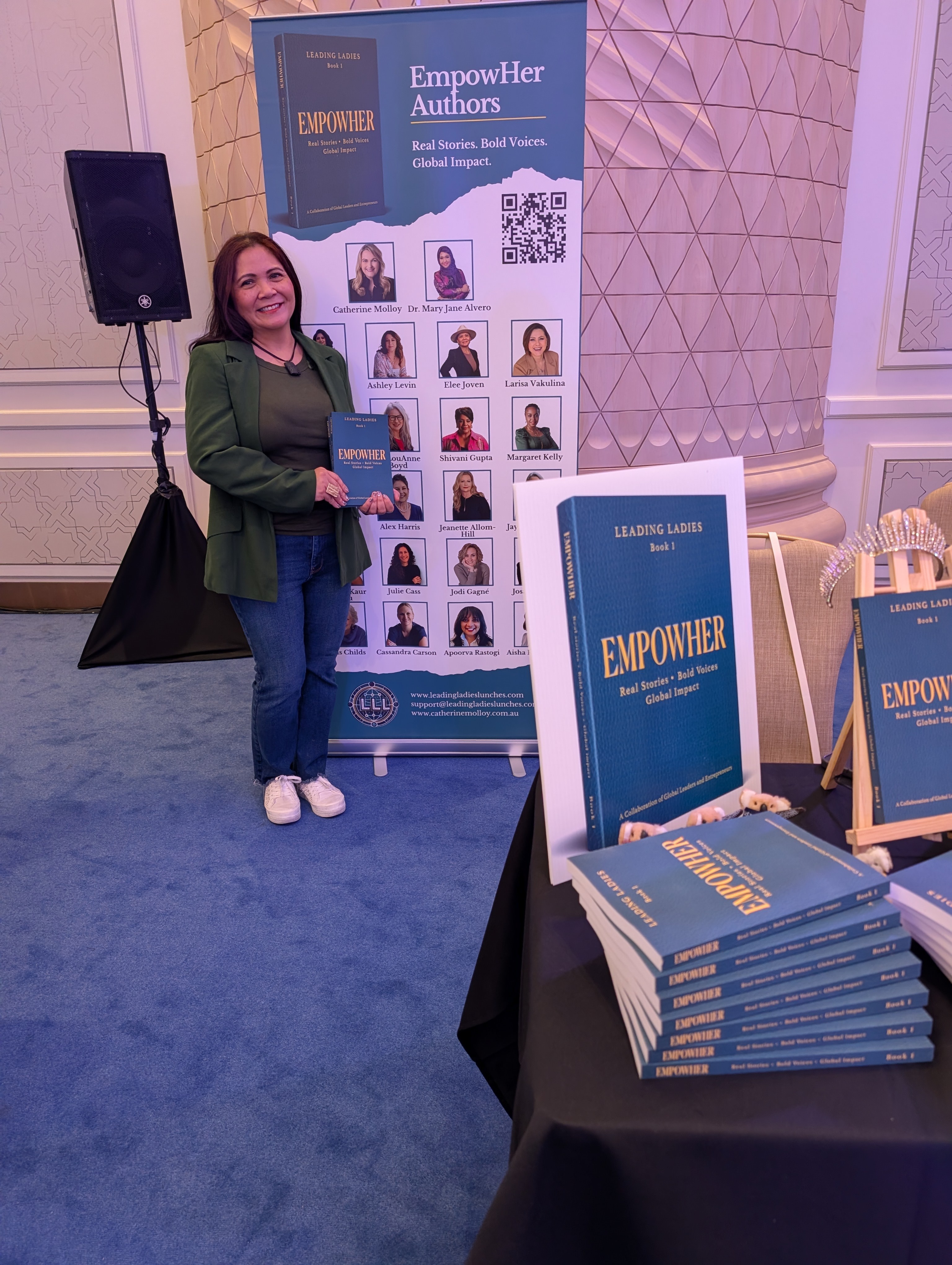 Woman standing next to a display banner at a book event, holding a book, with a table of stacked books nearby.