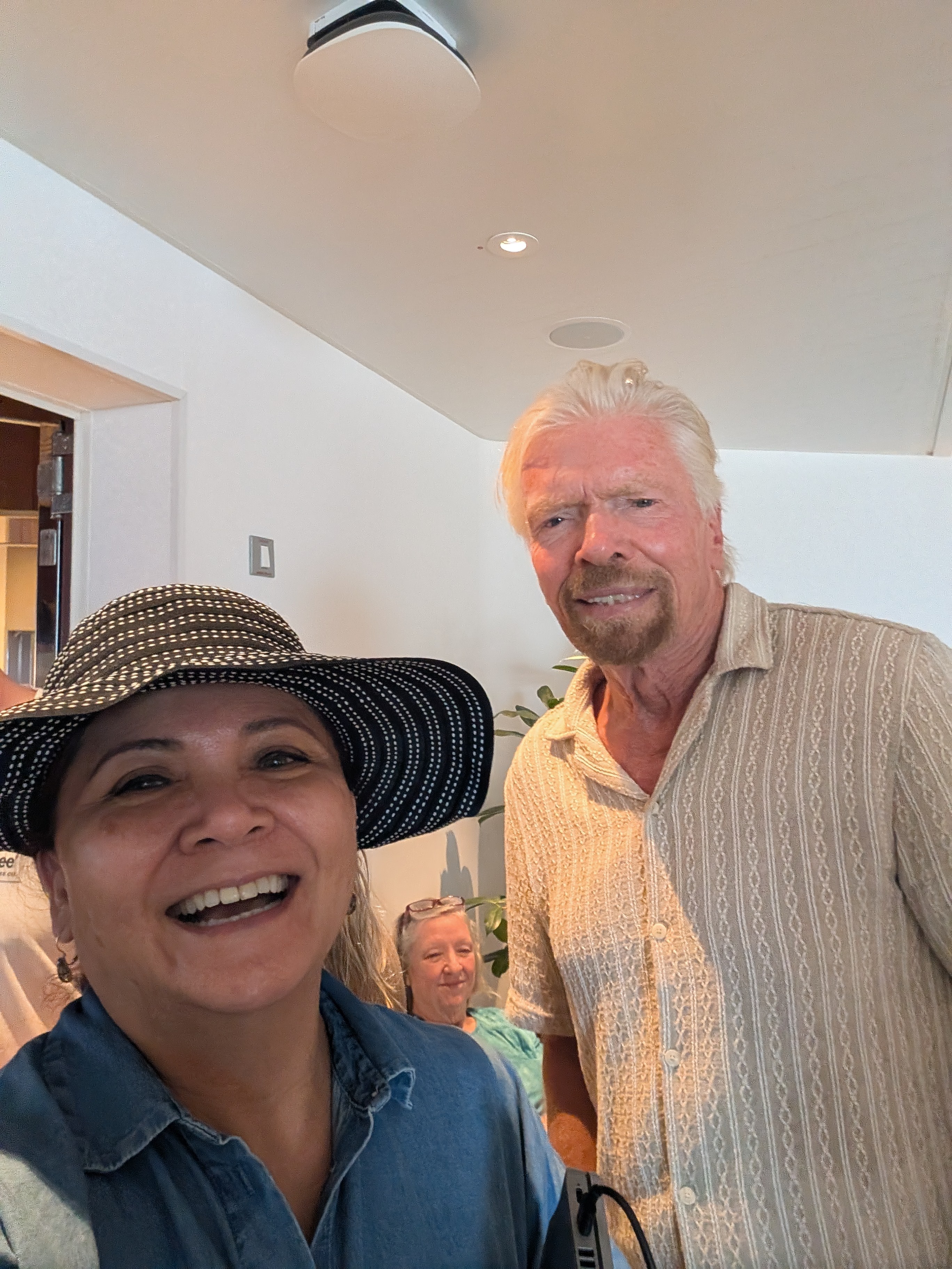 Two smiling people, a woman wearing a hat and a man with white hair, indoors with a white ceiling and light fixture.