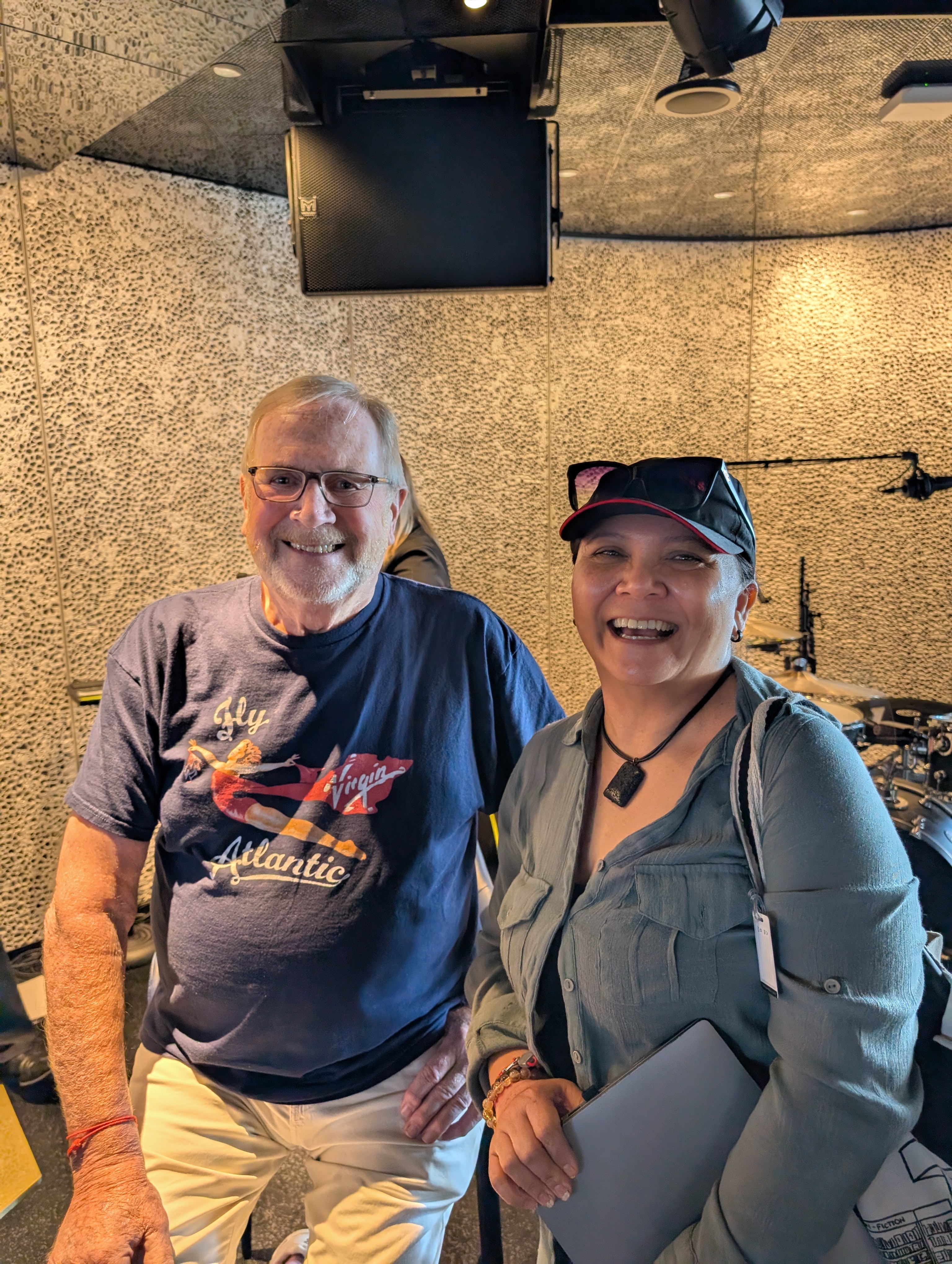 Two smiling people standing indoors, one wearing a navy T-shirt and the other in a gray jacket with a cap.
