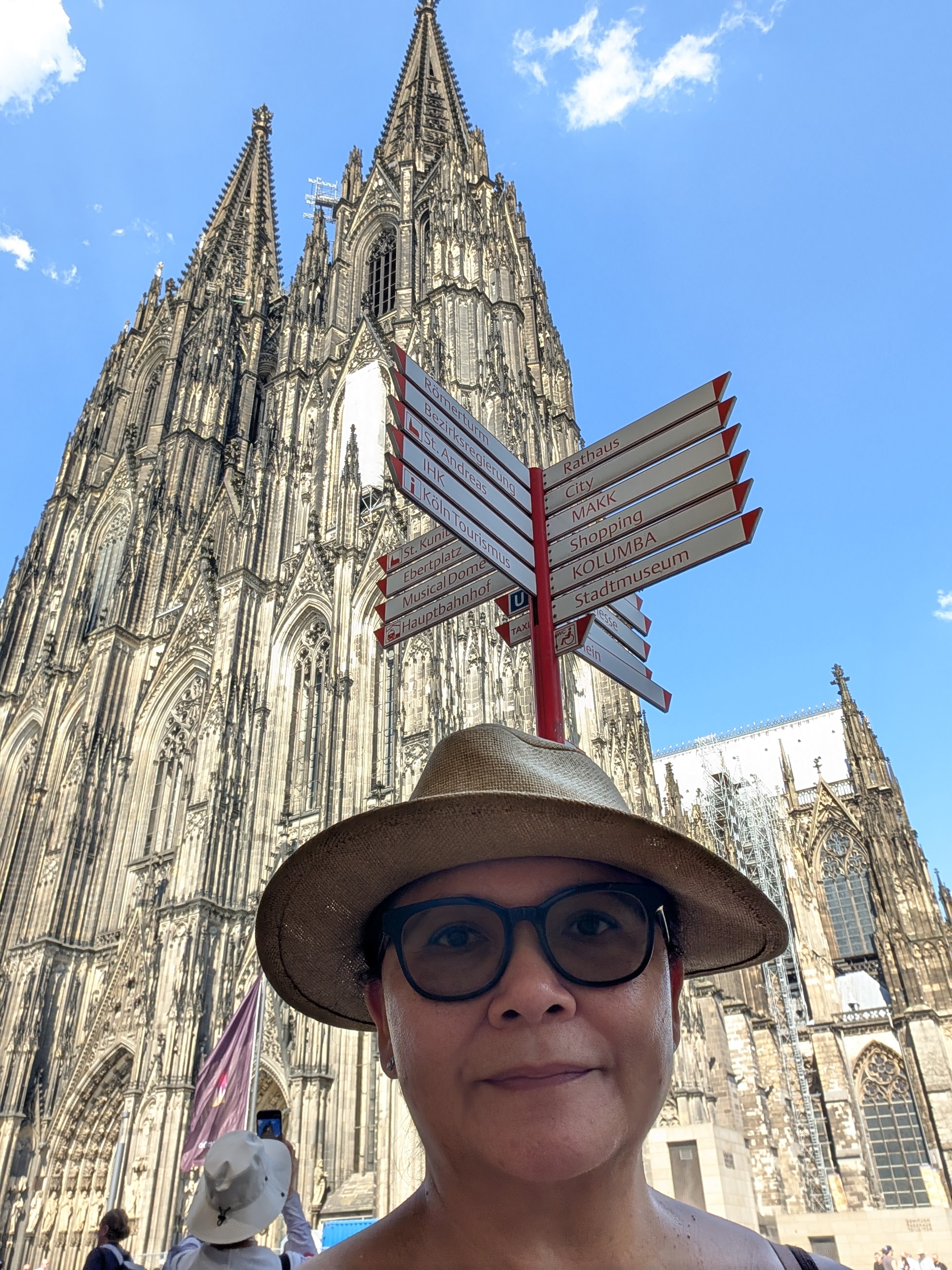 Woman wearing glasses and a hat in front of a Gothic cathedral with a signpost, blue sky background.