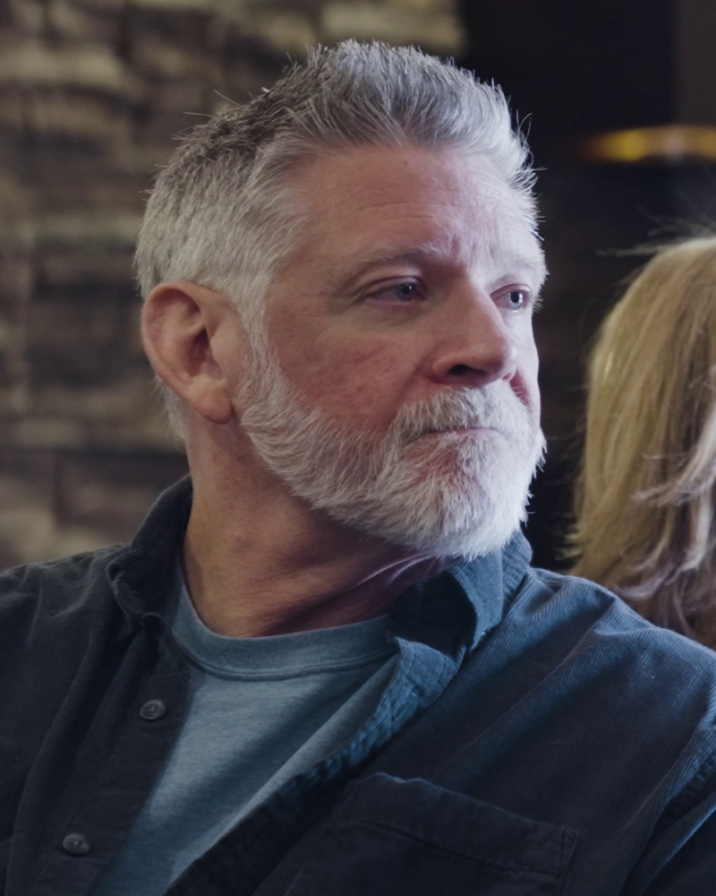 A middle-aged man with gray hair and a beard, wearing a dark shirt, looking to the side in an indoor setting.