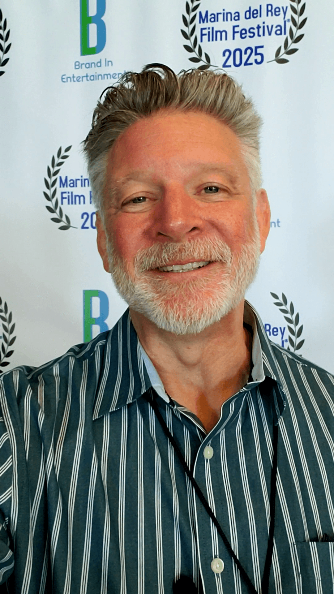 Smiling man with gray hair and beard wearing a striped shirt, standing in front of a festival backdrop.
