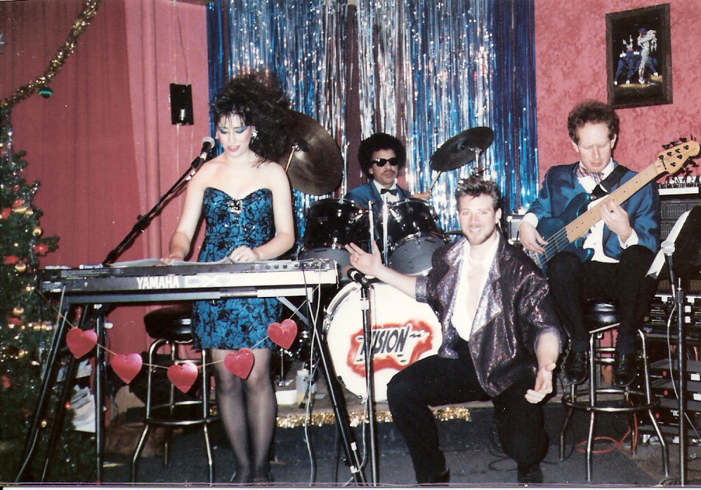 Four musicians perform on stage with pink curtains, a Christmas tree, and a shiny blue backdrop, including a woman at keyboard and three men with instruments.