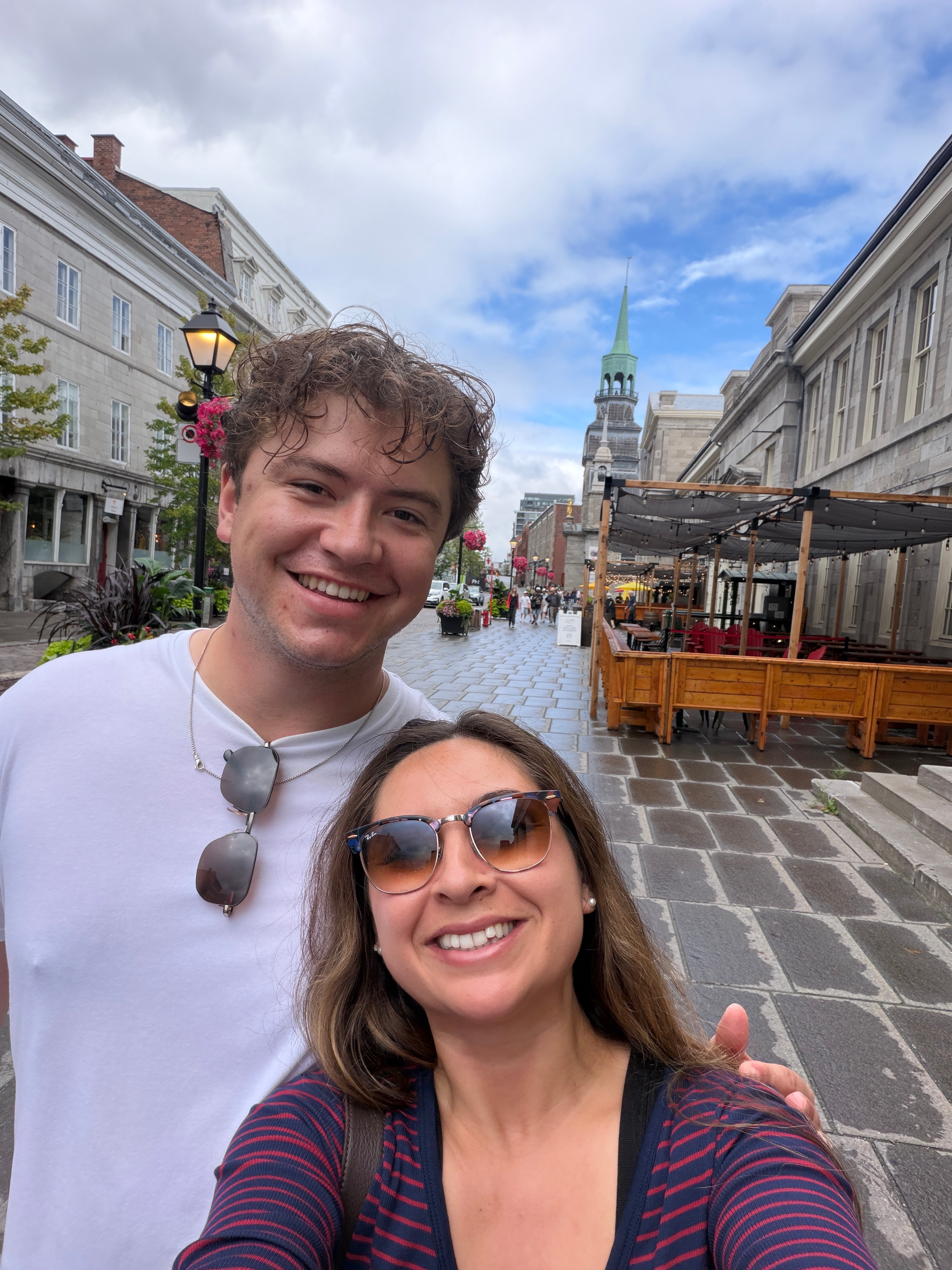 Two smiling people, a man and a woman, pose on a cobblestone street with historic buildings and a tower in the background.
